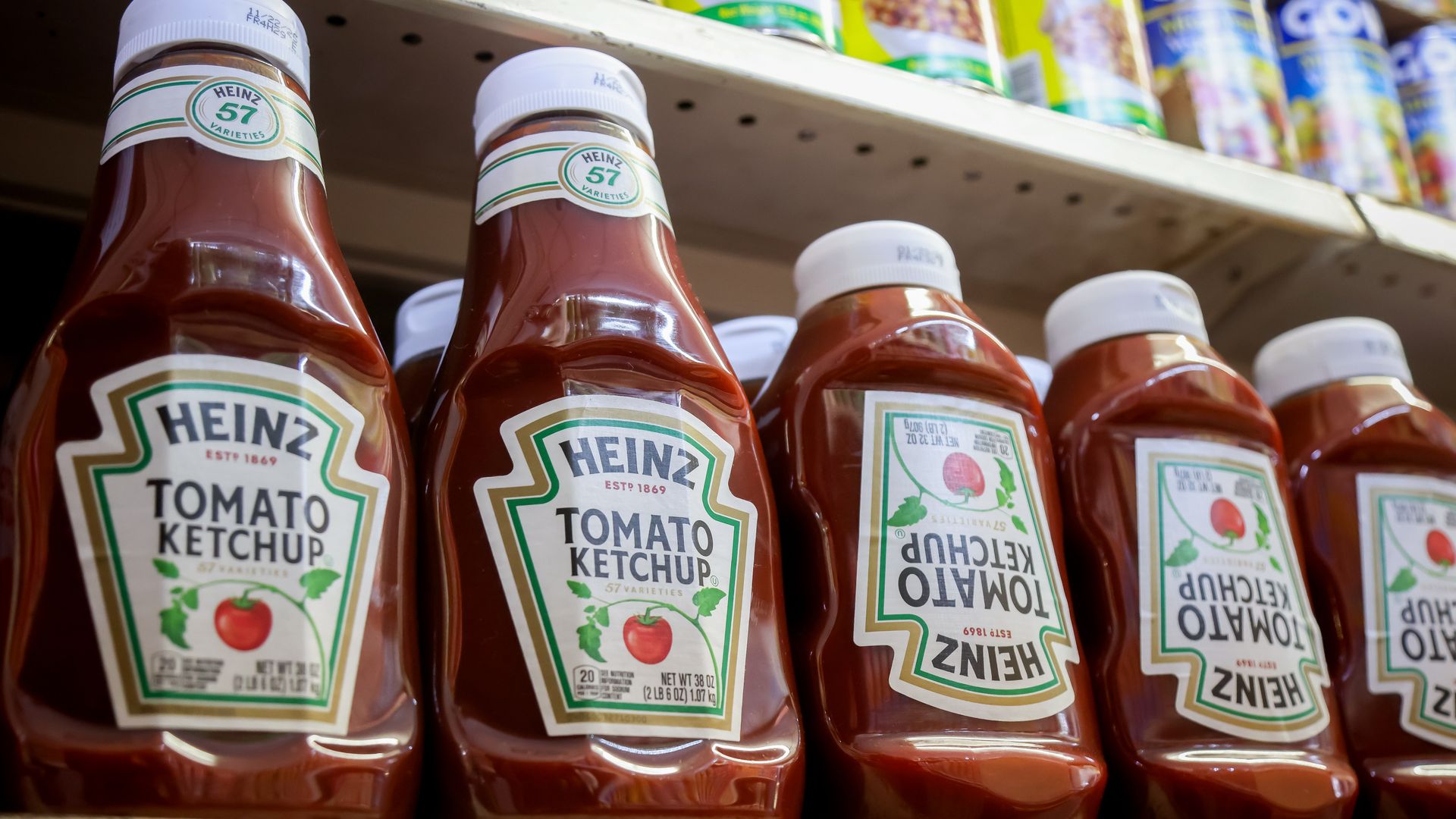 Row of Heinz Tomato Ketchup bottles with white caps on a grocery store shelf, some labels facing front and some upside down, with other canned goods on the shelf above.