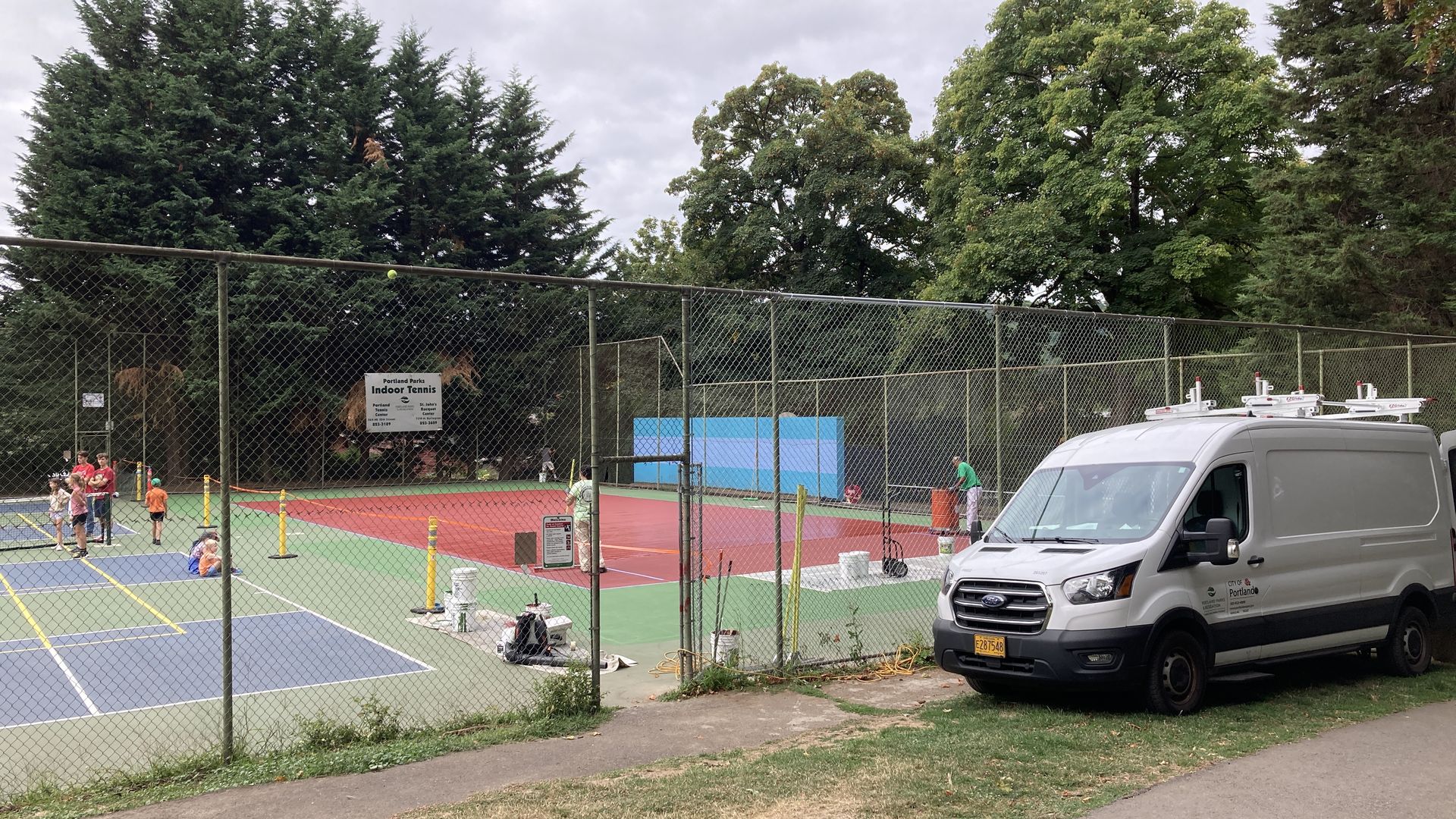 Two men applying red paint on a outdoor racquetball court, covering up the blue, white and green markings of a pickleball court.
