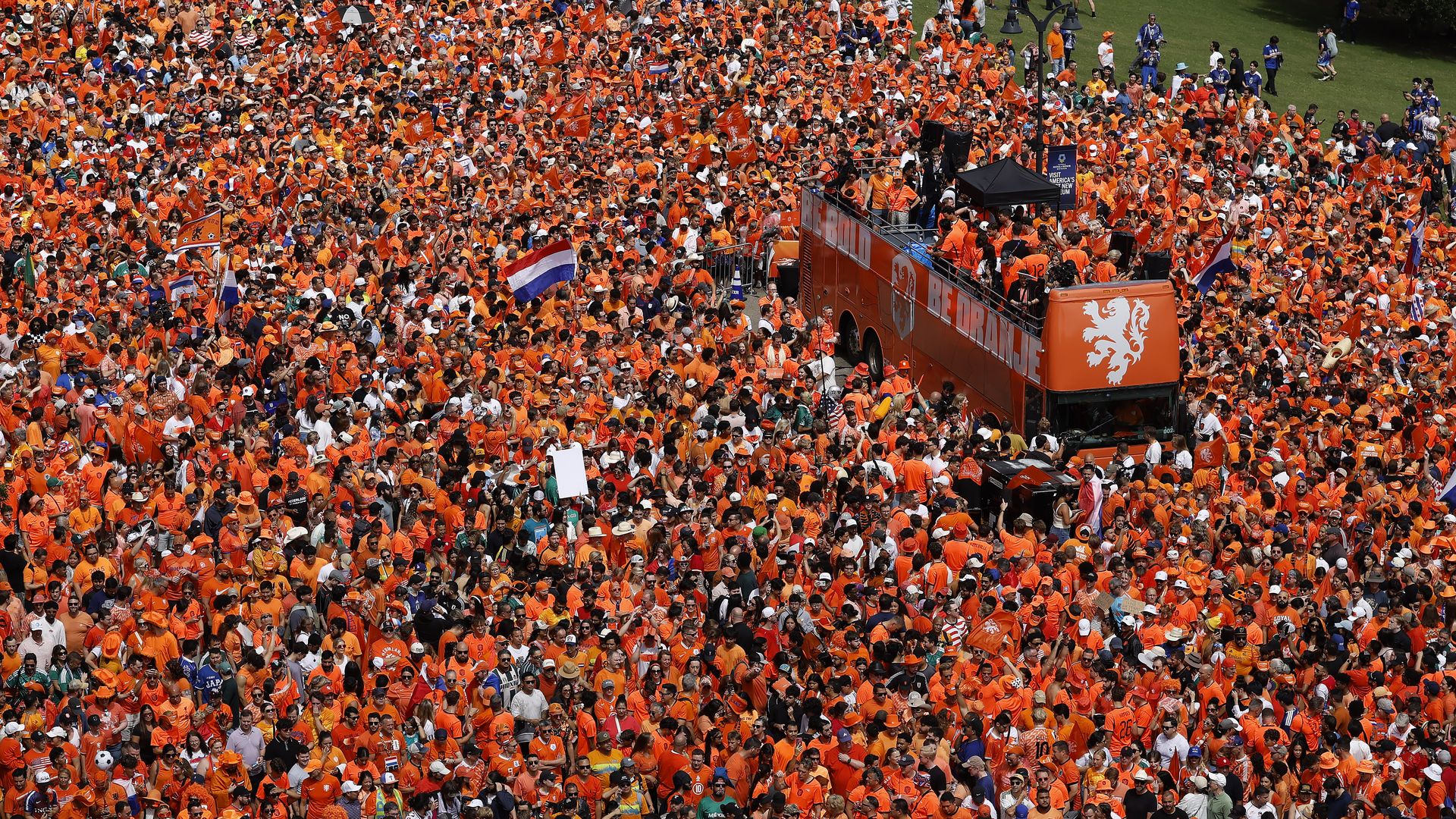 Overhead view of a massive crowd in orange shirts at a rally, waving orange and Dutch flags. An orange tour bus sits amid the sea of people and stage equipment.