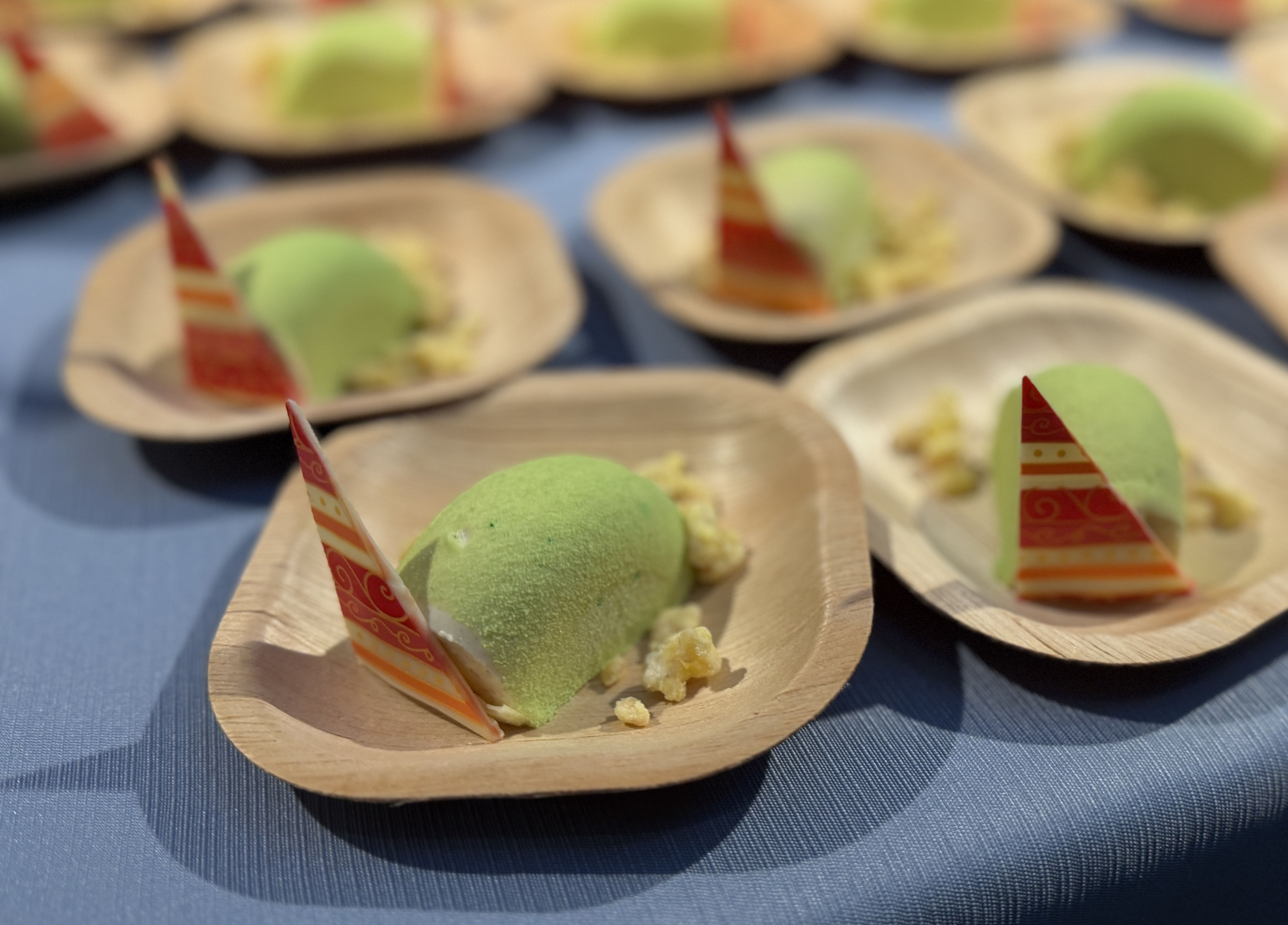 Blurry scene of small green dessert domes on boat-shaped plates, each with a red-striped decorative flag, arranged on a blue tablecloth.
