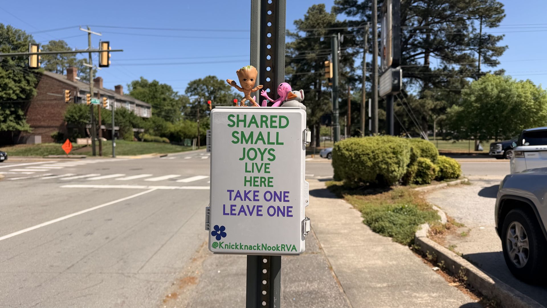 Street scene with a pole-mounted sign reading "SHARED SMALL JOYS LIVE HERE TAKE ONE LEAVE ONE"; two toys, Groot and a pink flamingo, perch on top. Clear blue sky, trees, and vehicles in the background.
