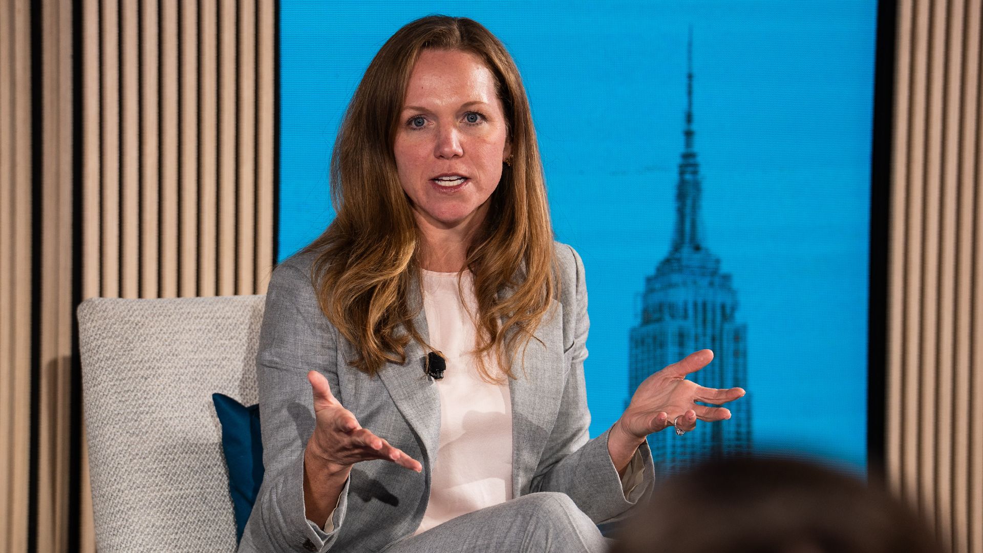 Woman with long brown hair in a light gray suit and pale pink blouse speaks and gestures with hands while seated on a light gray chair with blue background featuring a blurred tall building.