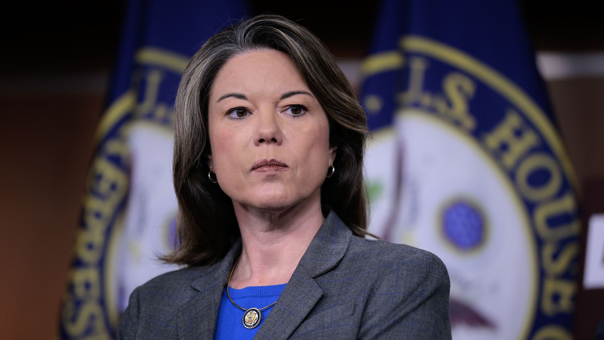 Woman with brown hair wearing a blue shirt and gray blazer standing in front of blurred U.S. House of Representatives flags.