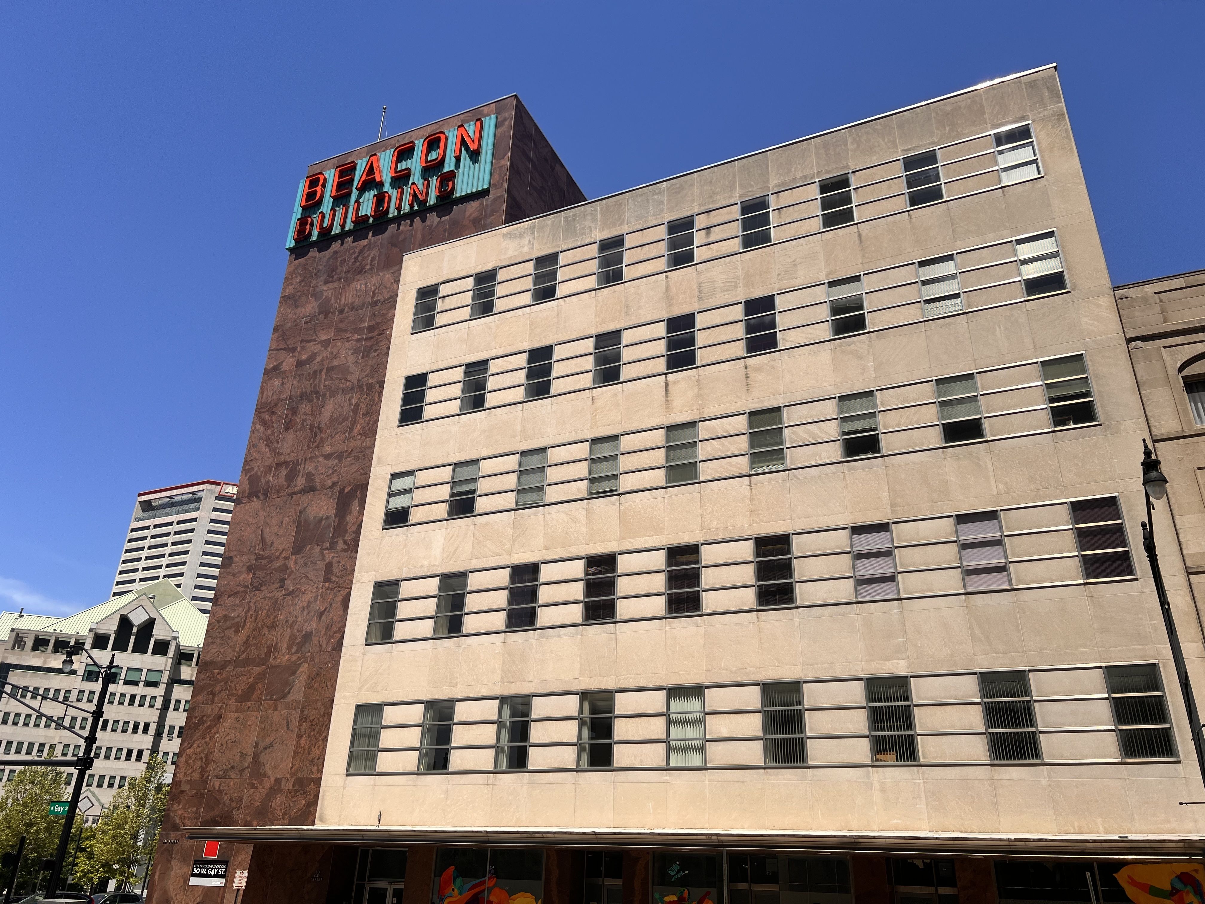 A tall beige and brown office building against a clear blue sky with a turquoise sign reading BEACON BUILDING in red letters on the left; many rectangular windows form a grid.