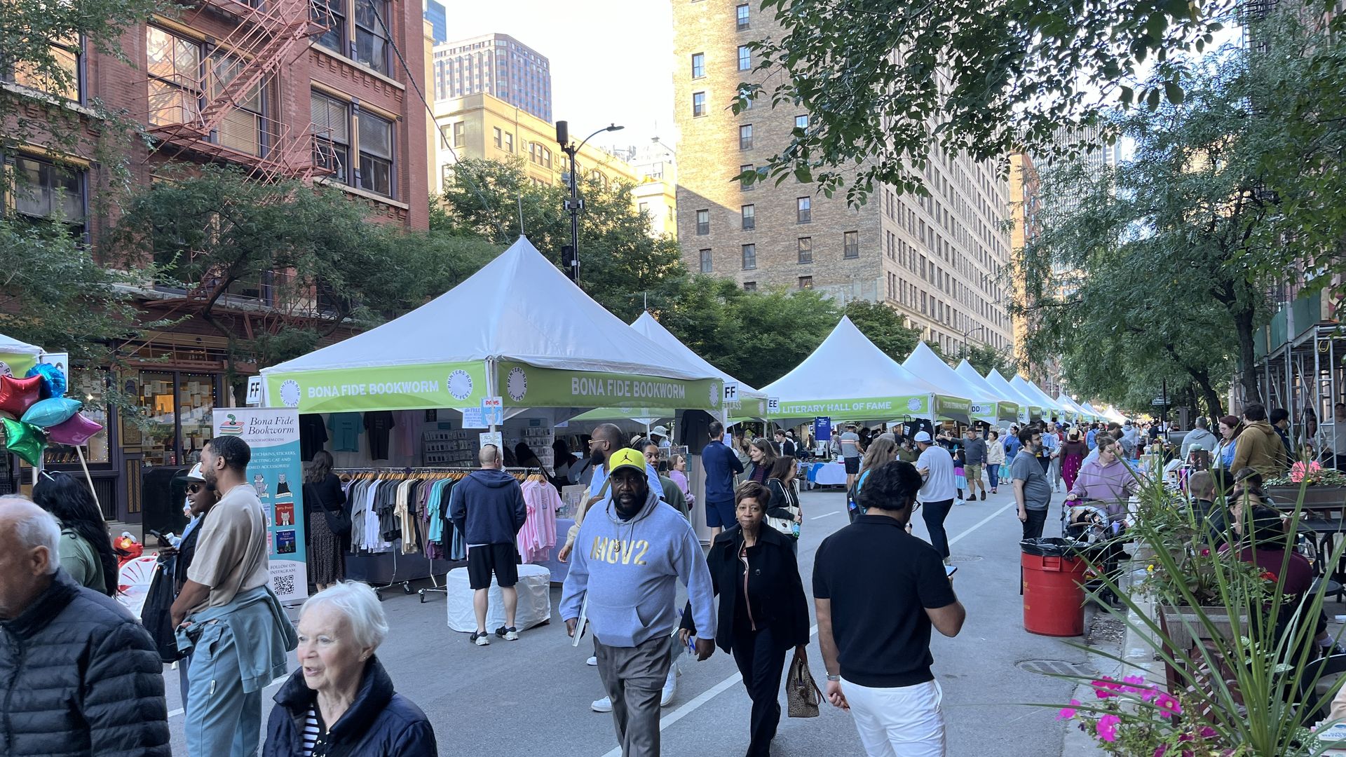 Outdoor street market with white tents labeled "Bona Fide Bookworm" and "Literary Hall of Fame," people walking and browsing, city buildings and trees in the background on a sunny day.