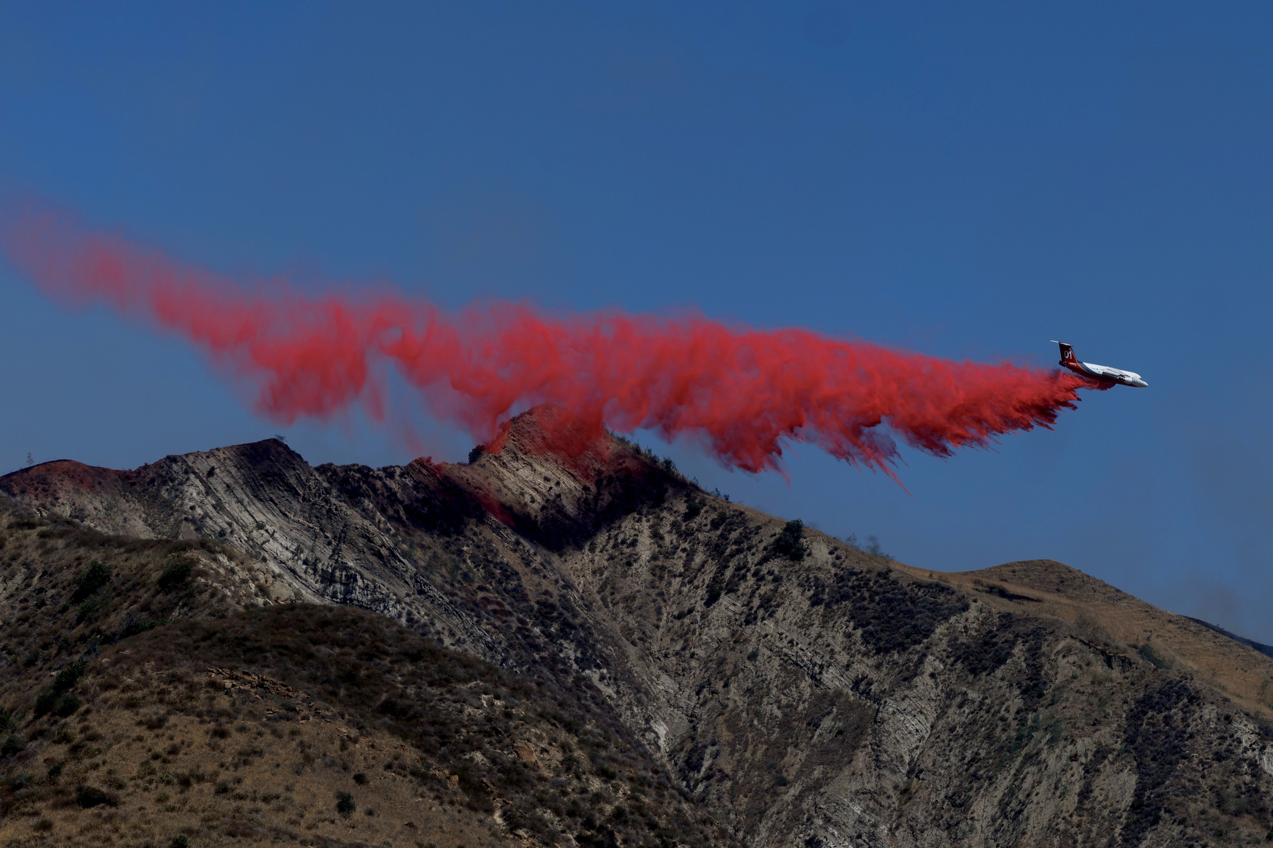 Firefighting airplane releasing a large plume of bright red fire retardant over dry, rugged mountain terrain under a clear blue sky.