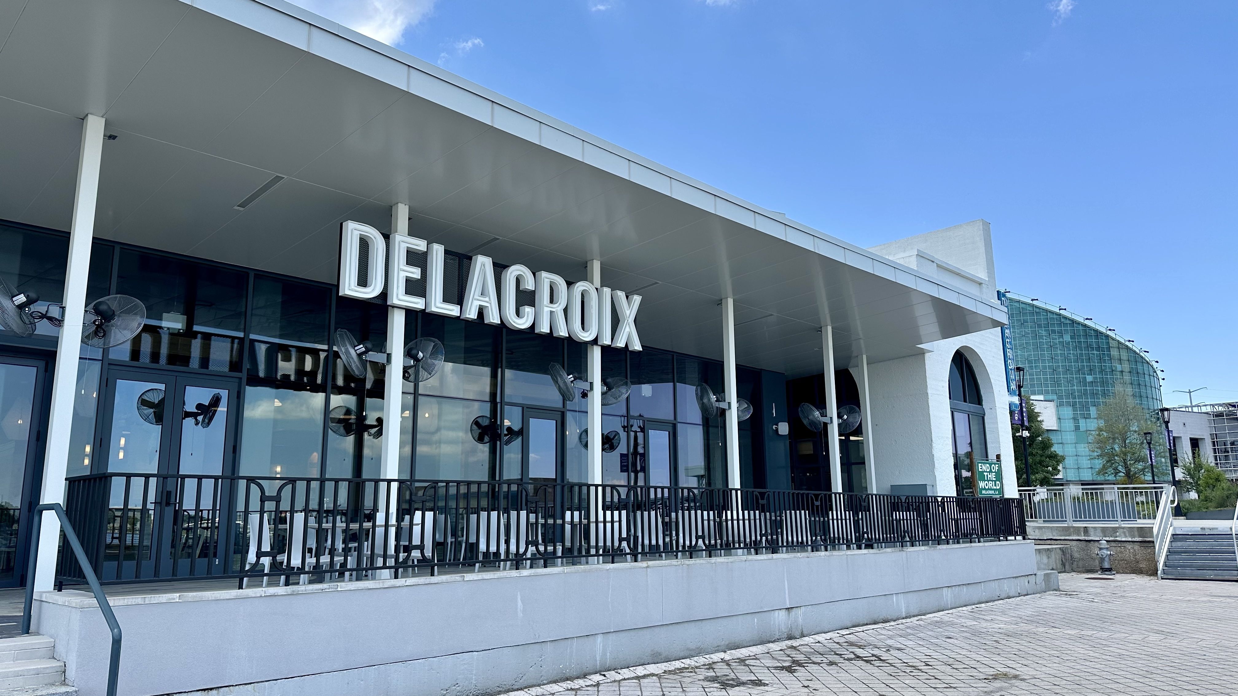 Exterior street view of Delacroix restaurant with large white letters on a modern glass building and a clear blue sky above, featuring black railings and outdoor fans.