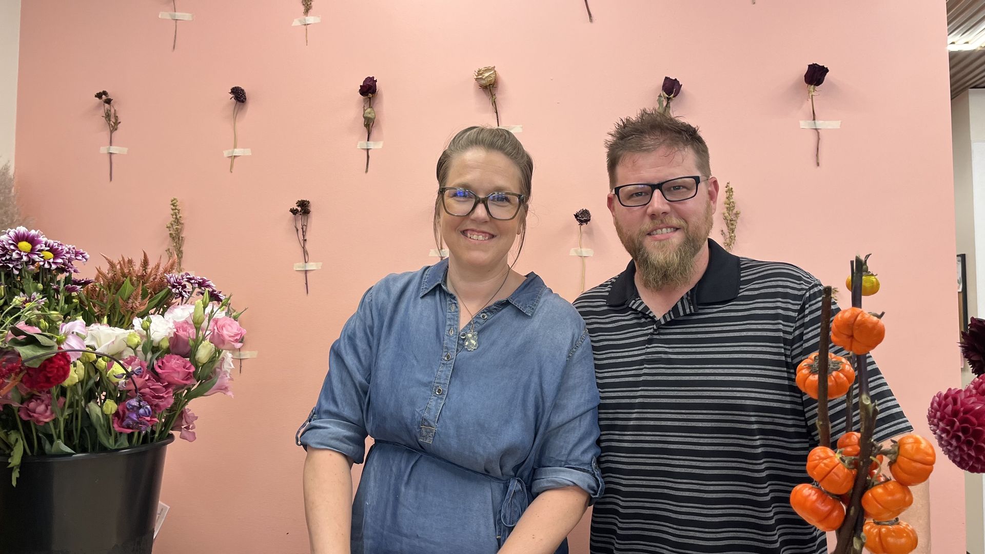 Stacey and Brandt stand in front of a pink wall decorated with dried flowers taped in a grid. Colorful fresh flowers and small orange gourds are in foreground.