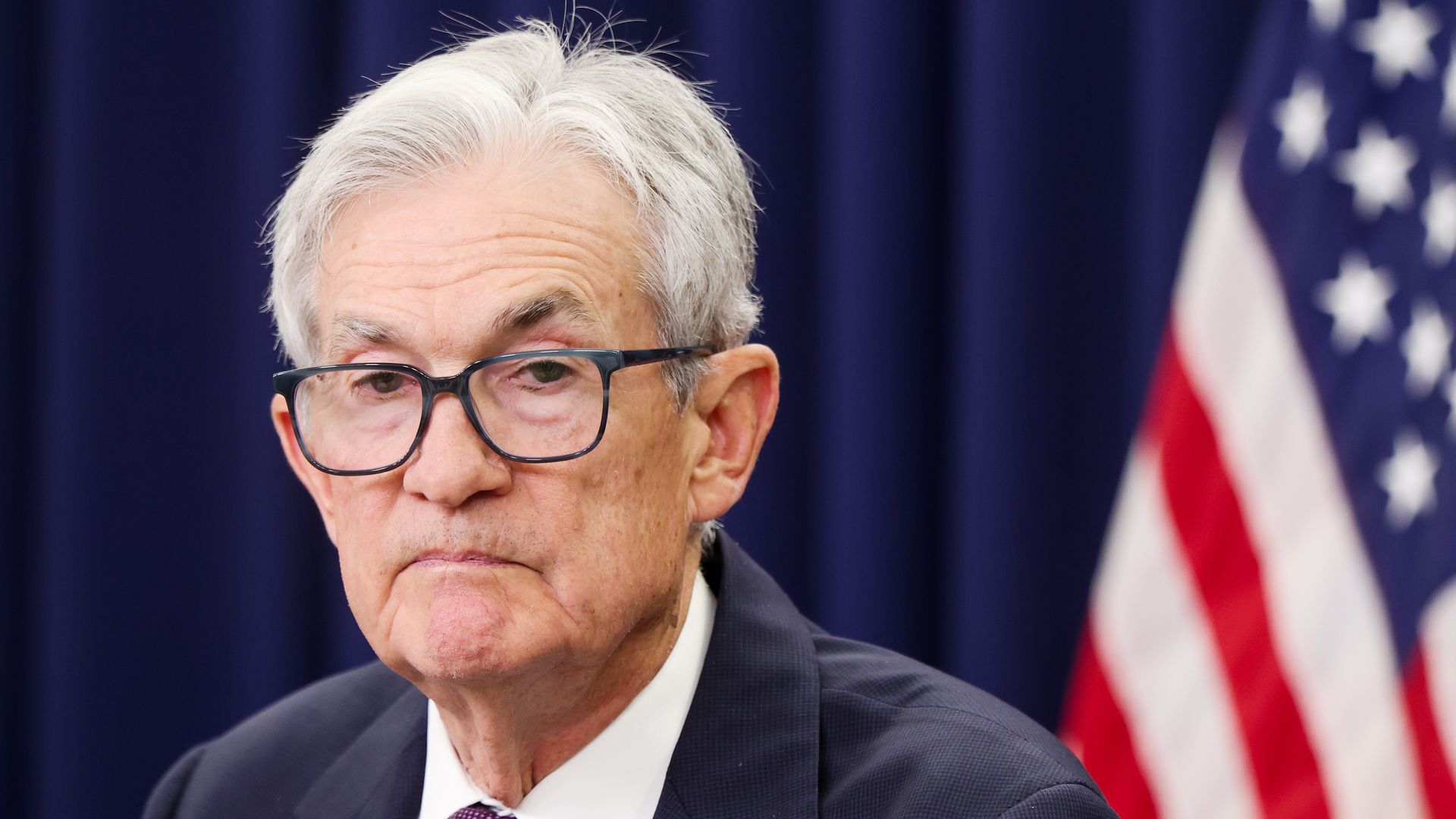 Close-up of a man with gray hair and glasses in a dark suit and purple tie, against a dark blue backdrop with part of the American flag in the background.