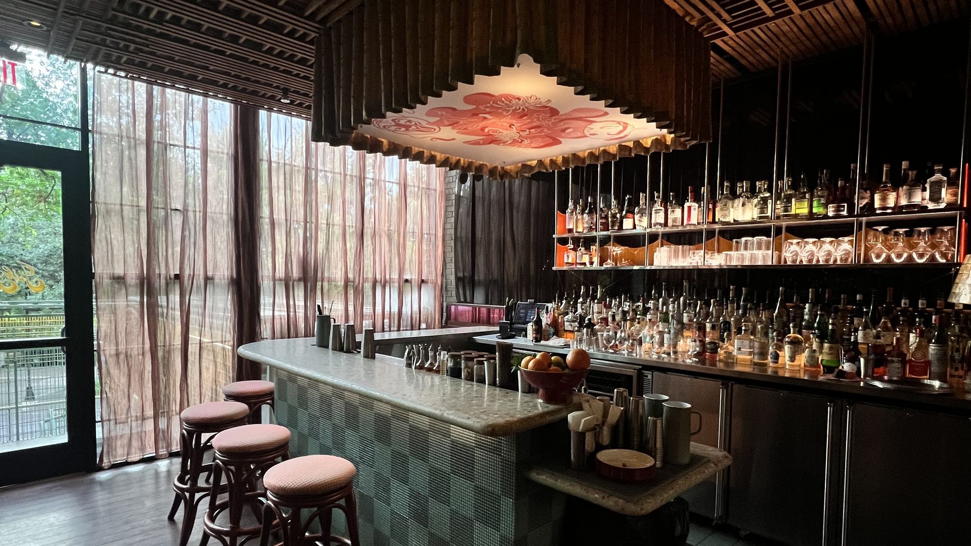 Dimly lit bar with a dark wood slat ceiling and a large square chandelier-like fixture showing a red floral pattern. A tiled bar counter with stools and shelves of liquor bottles along the back wall.