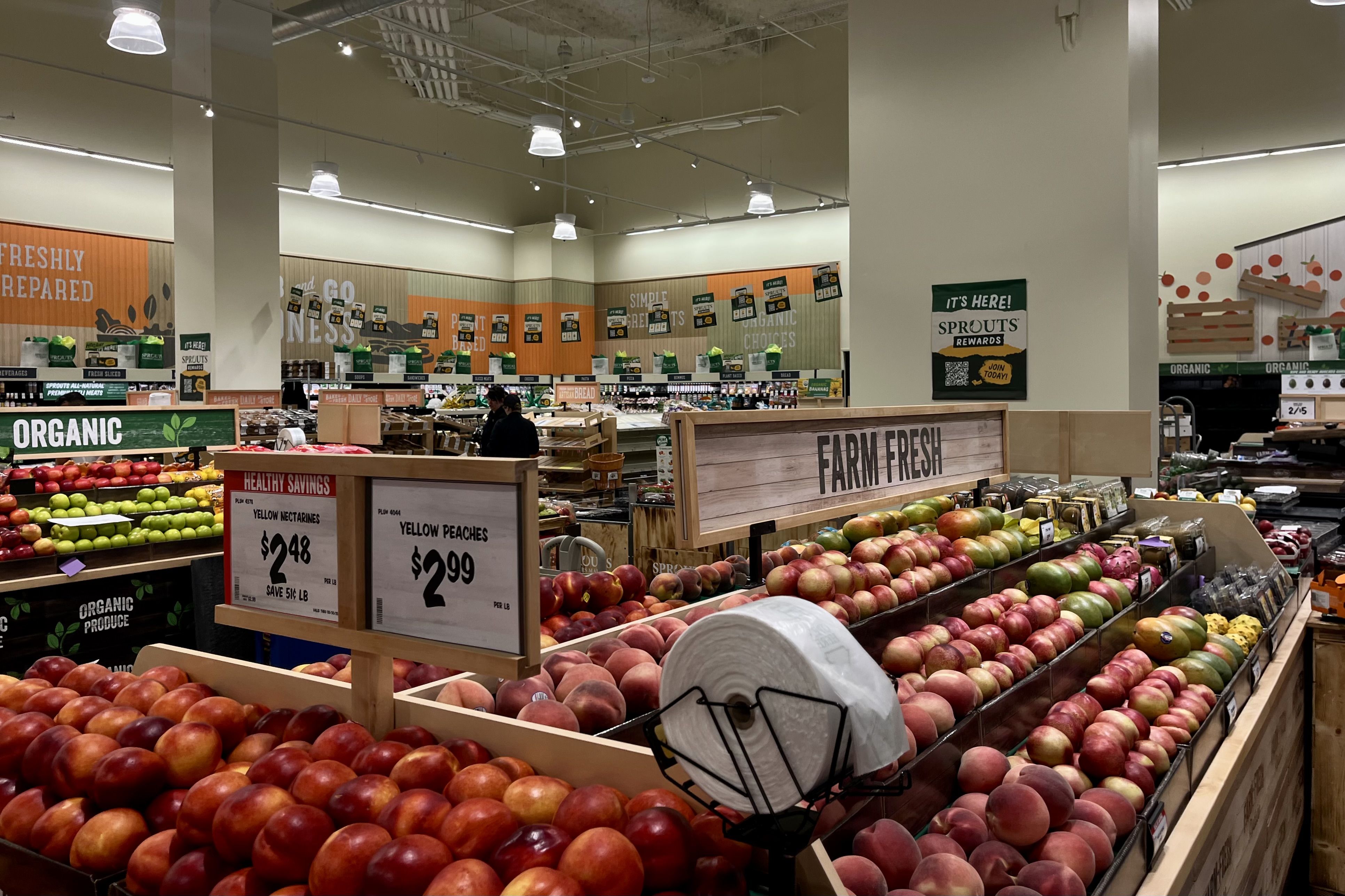 Grocery store display with assorted fresh vegetables including carrots, artichokes, asparagus, cucumbers, yellow bell peppers, red bell peppers, eggplants, and green bell peppers under a "FARM FRESH" sign.