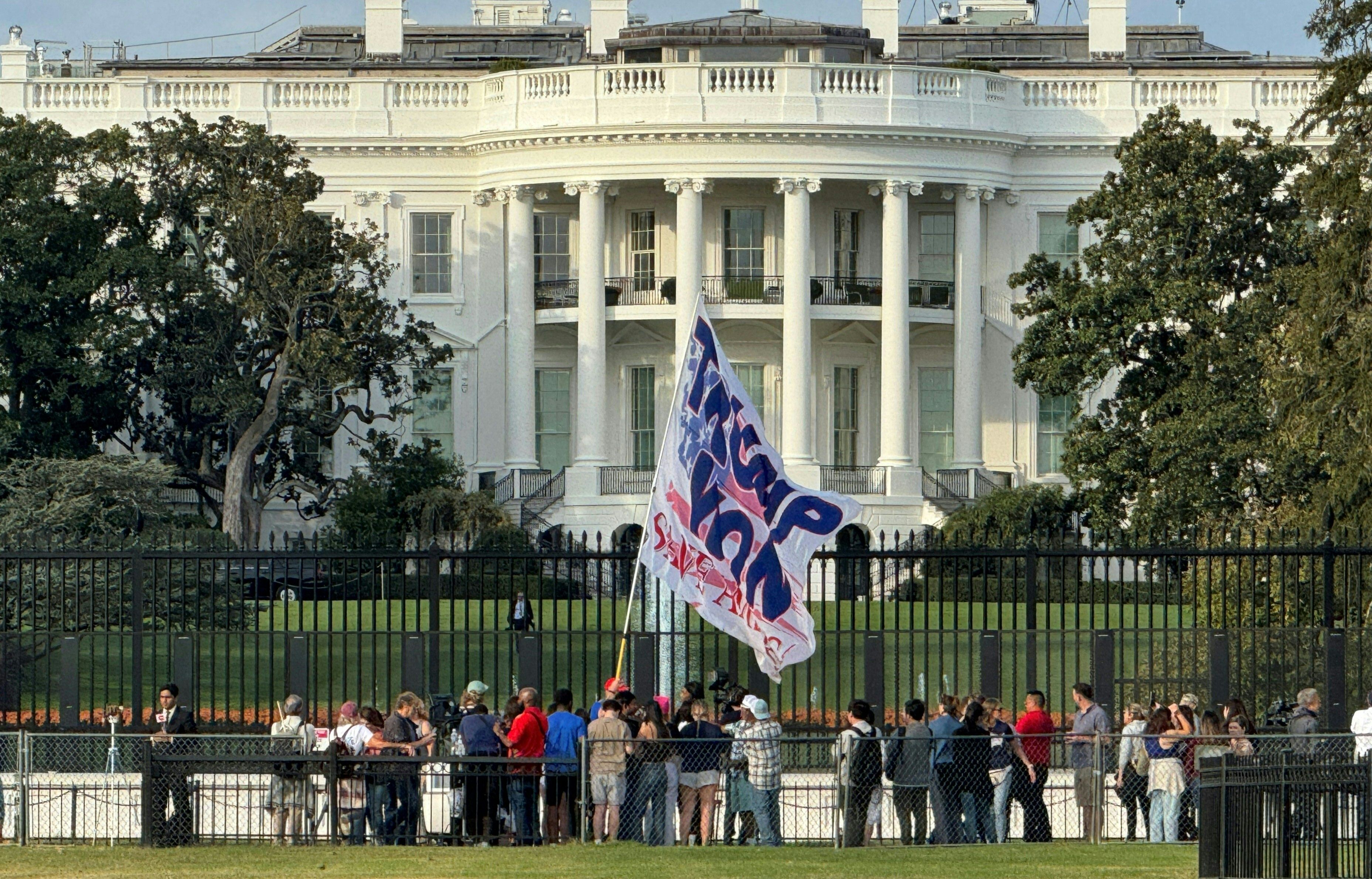 A crowd featuring a person waving a "Trump Won" stands outside the White House.