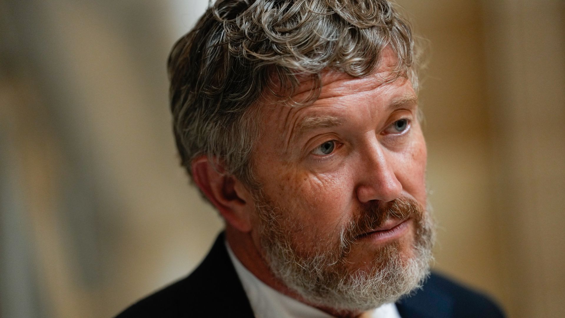 Close-up of a middle-aged man with curly gray hair and beard, wearing a white shirt, patterned tie, and dark suit jacket, looking thoughtfully to the side against a blurred beige background.