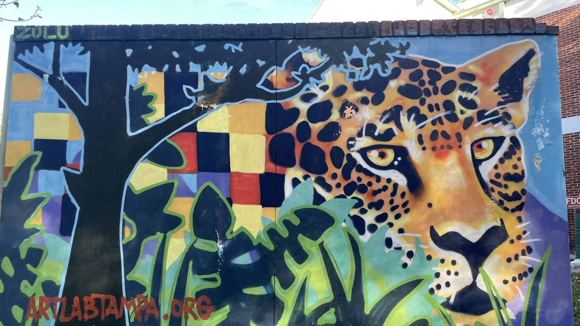 A mural of a leopard next to a tree and other plant life with a yellow, orange, red and purple checkered backdrop.