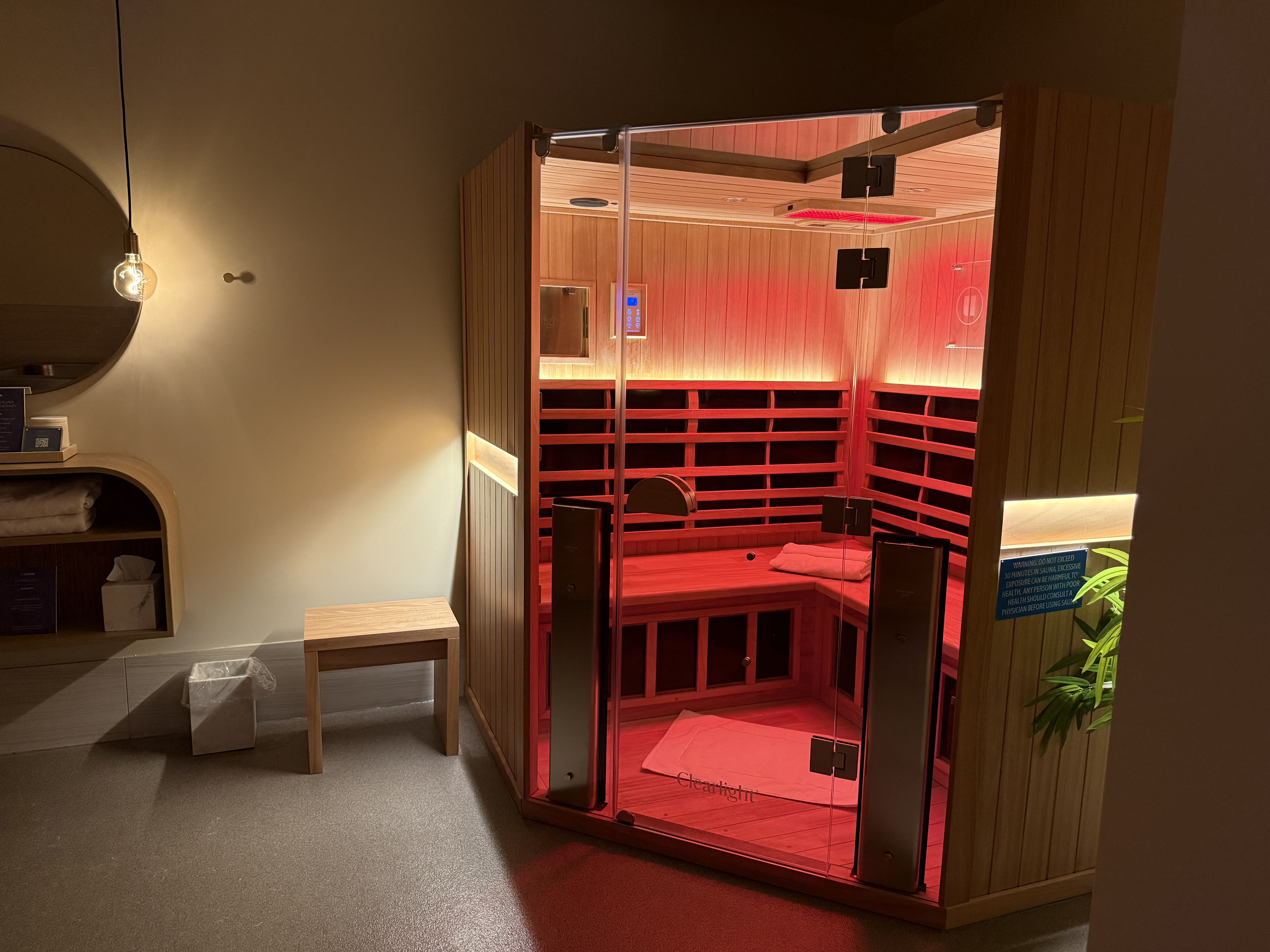 Modern wooden sauna room with red lighting, glass door, towel on bench, adjacent stool, mirror, shelf with towels, ambient warm lighting in cozy spa space.
