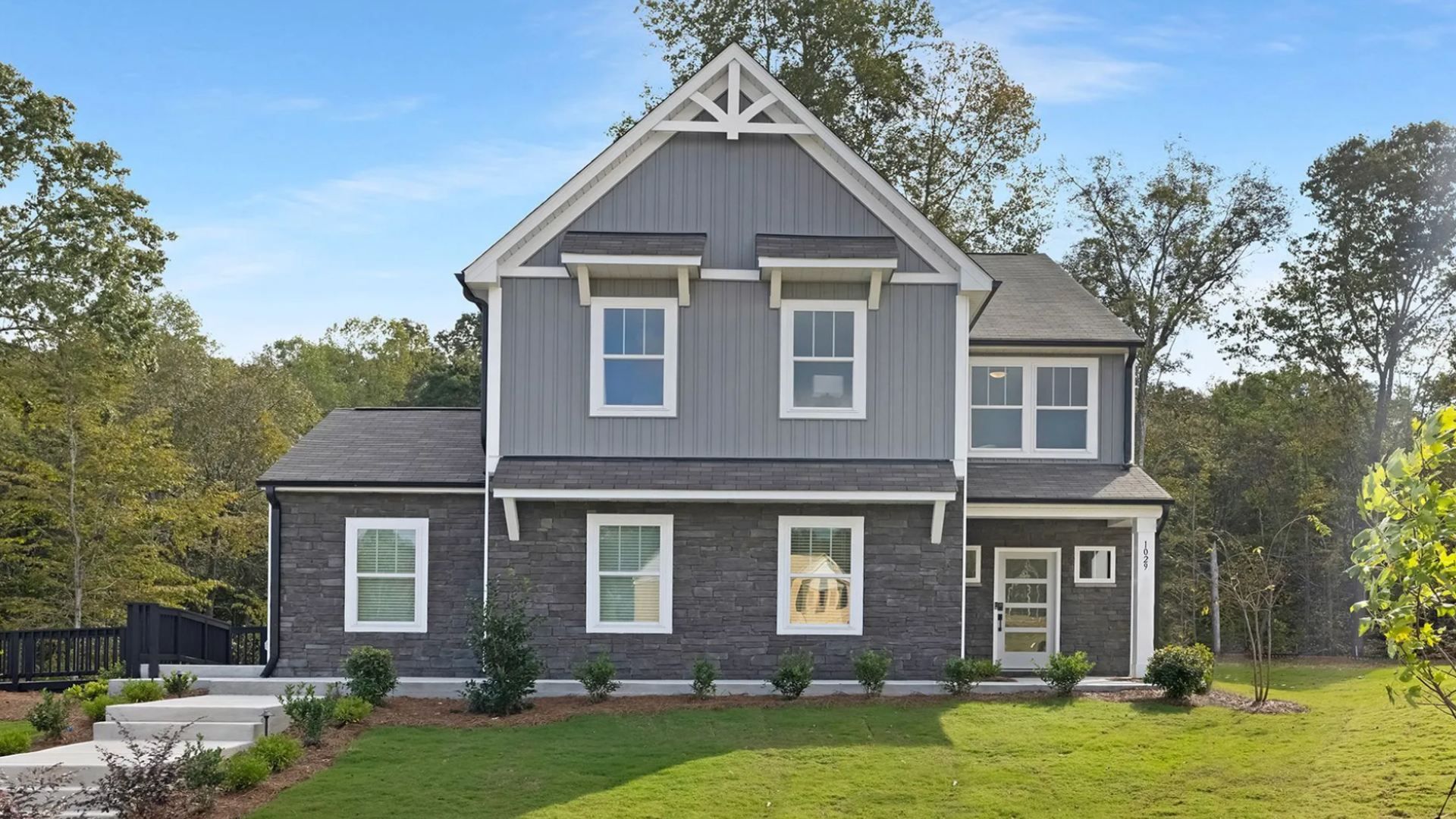 Two-story gray house with stone lower façade, white trim, and decorative gable. Front porch, multiple white-framed windows, manicured lawn, and trees against a blue sky.