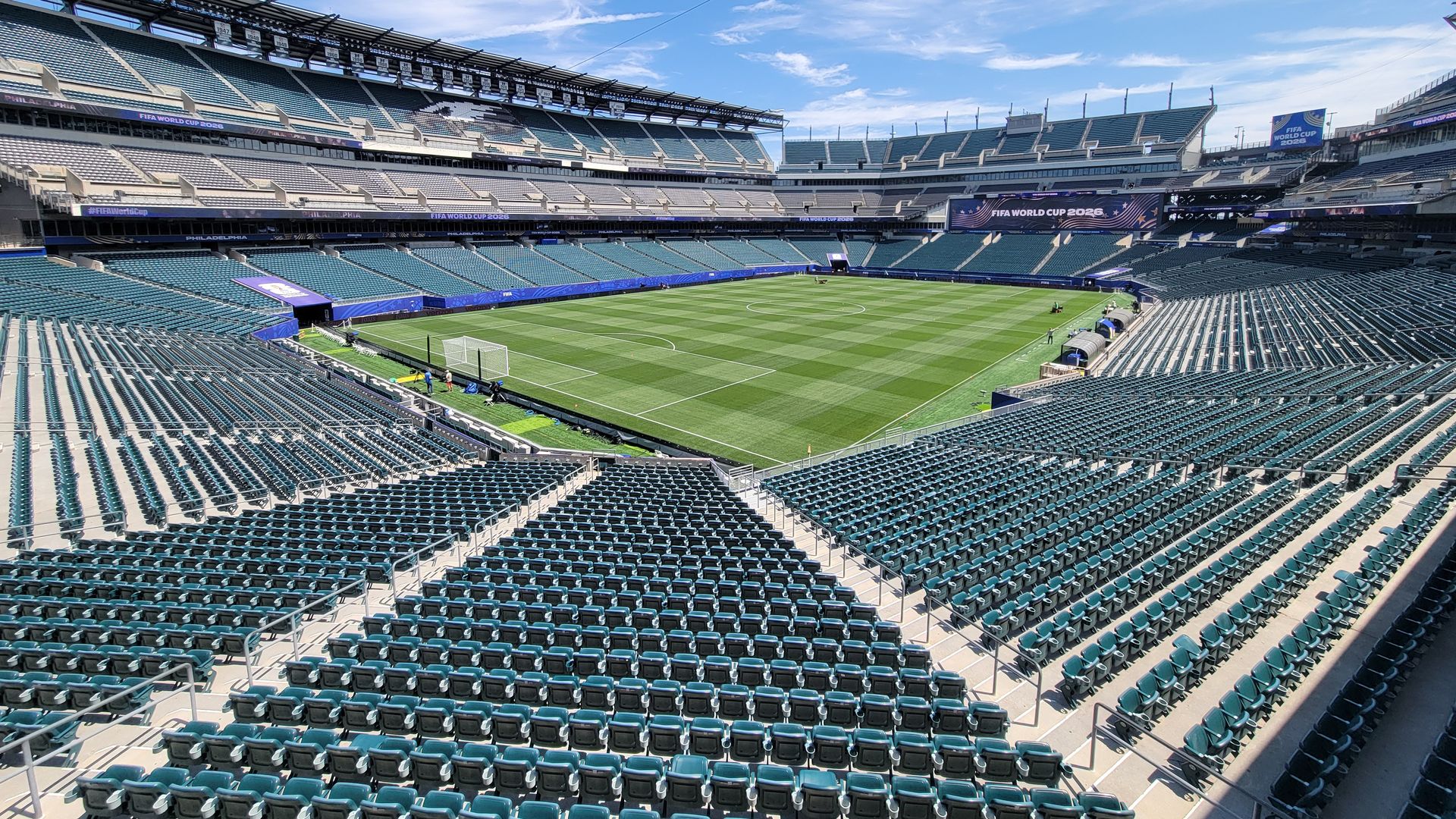 Empty outdoor soccer stadium with bright blue seats surrounding a green pitch; banners read FIFA World Cup 2026, and a clear blue sky overhead.