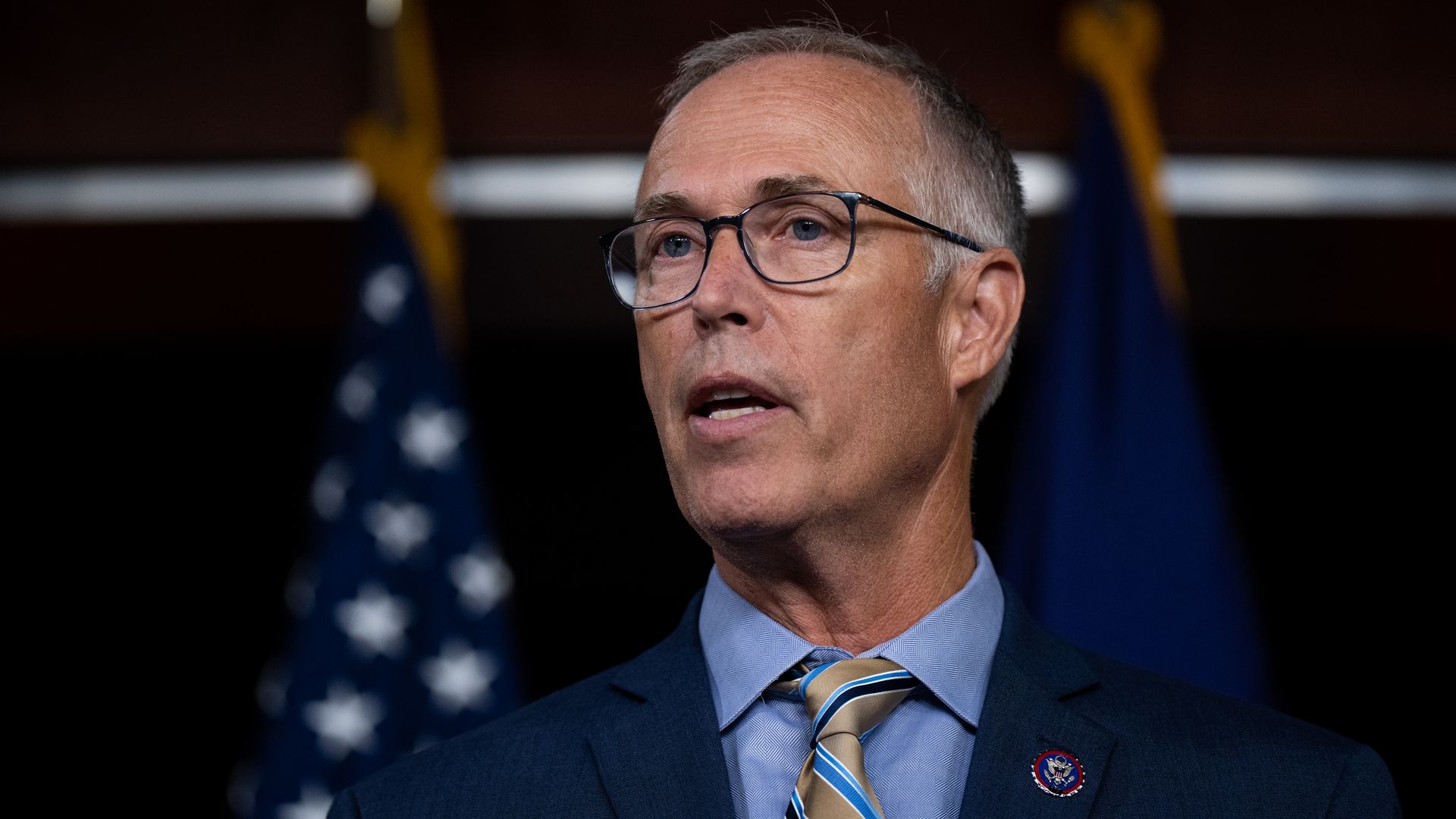 Rep. Jared Huffman, wearing a blue suit and speaking in front of American flags.