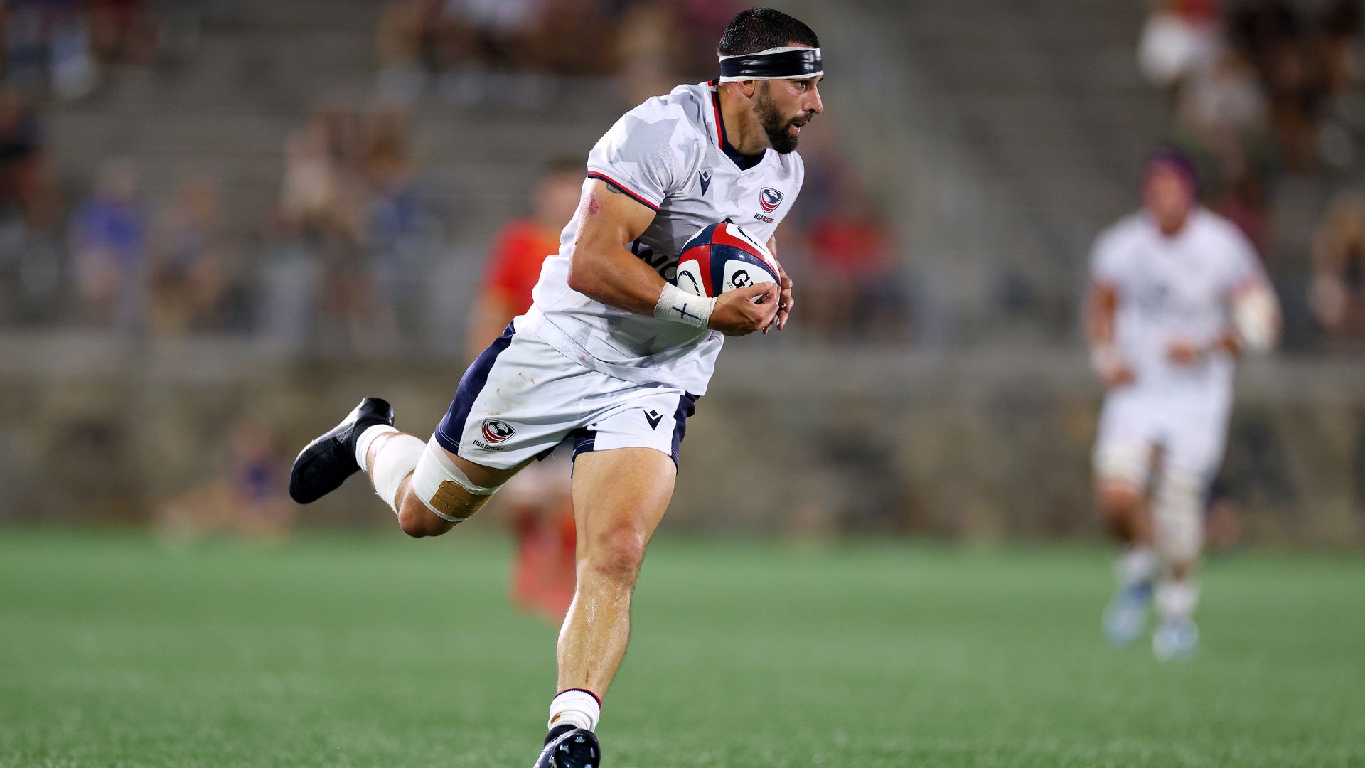 A rugby player dressed in a white uniform, blue sweatband, white socks and black cleats runs with red, white and blue rugby ball on a field. 