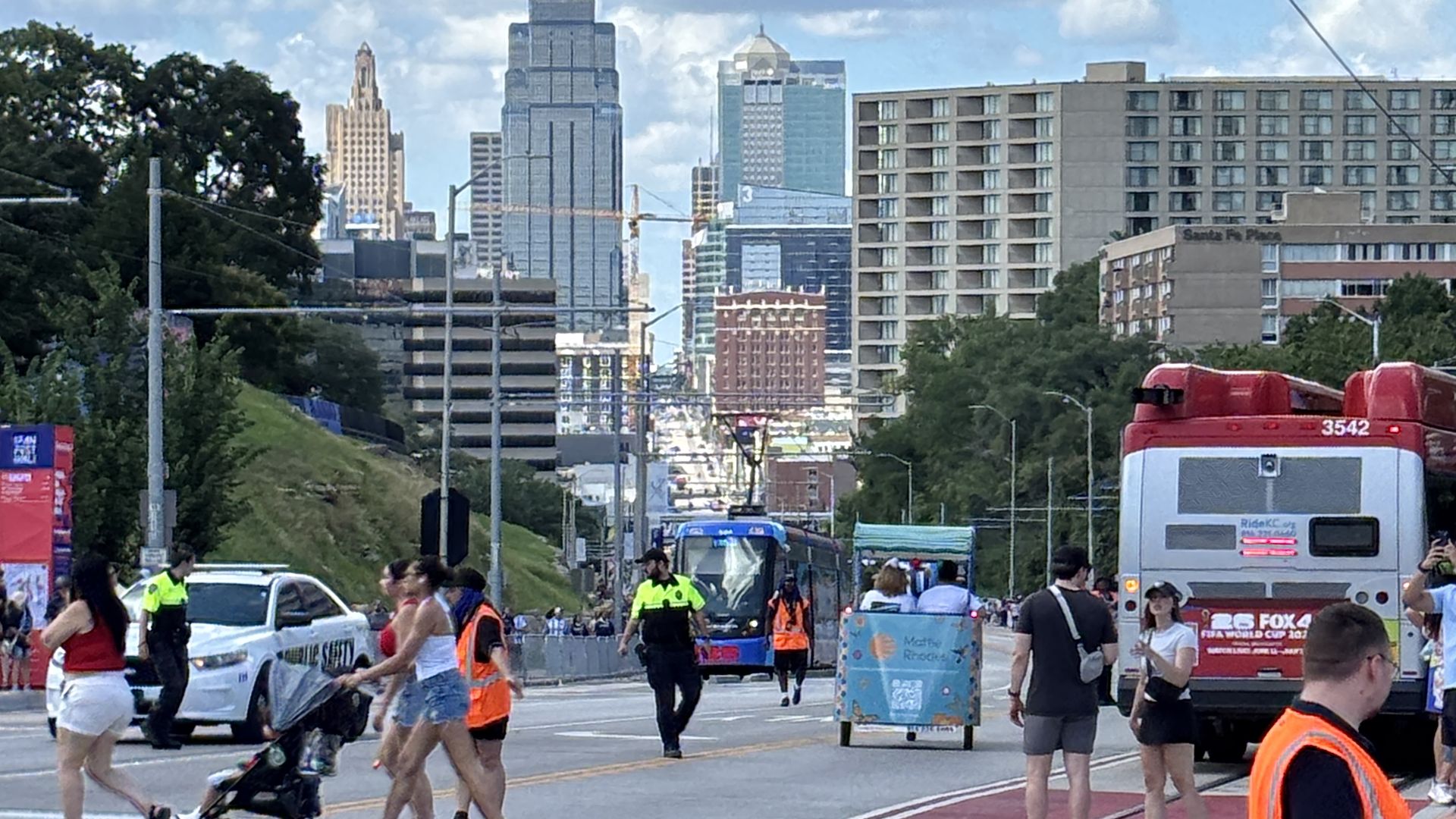 City street race scene with runners and volunteers in neon vests, a red bus at right, and a modern downtown skyline with cranes and tall glass buildings under a blue, cloudy sky.