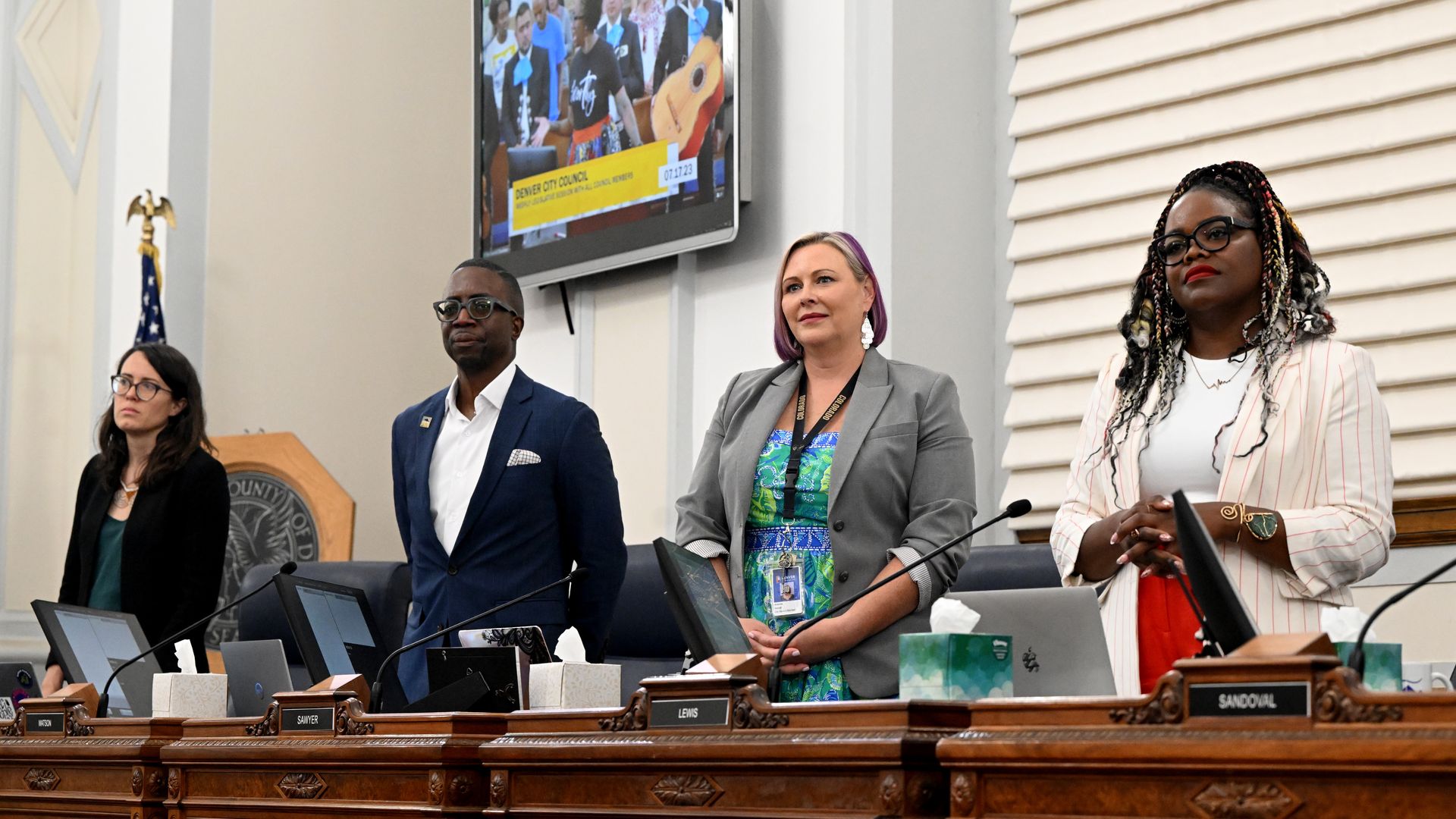 Four people rise in various poses in front of wooden desks inside a large chamber, wearing professional clothing.