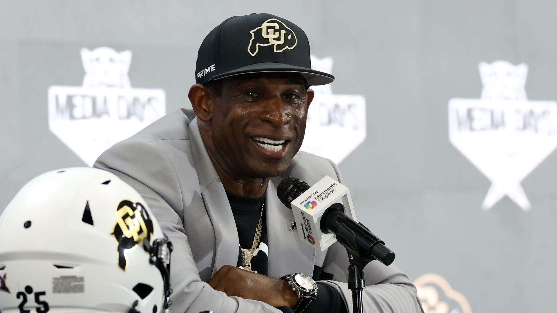 Head coach Deion Sanders of the Colorado Buffaloes sits at a press conference wearing a CU Buffs cap, with a helmet on the table next to him.