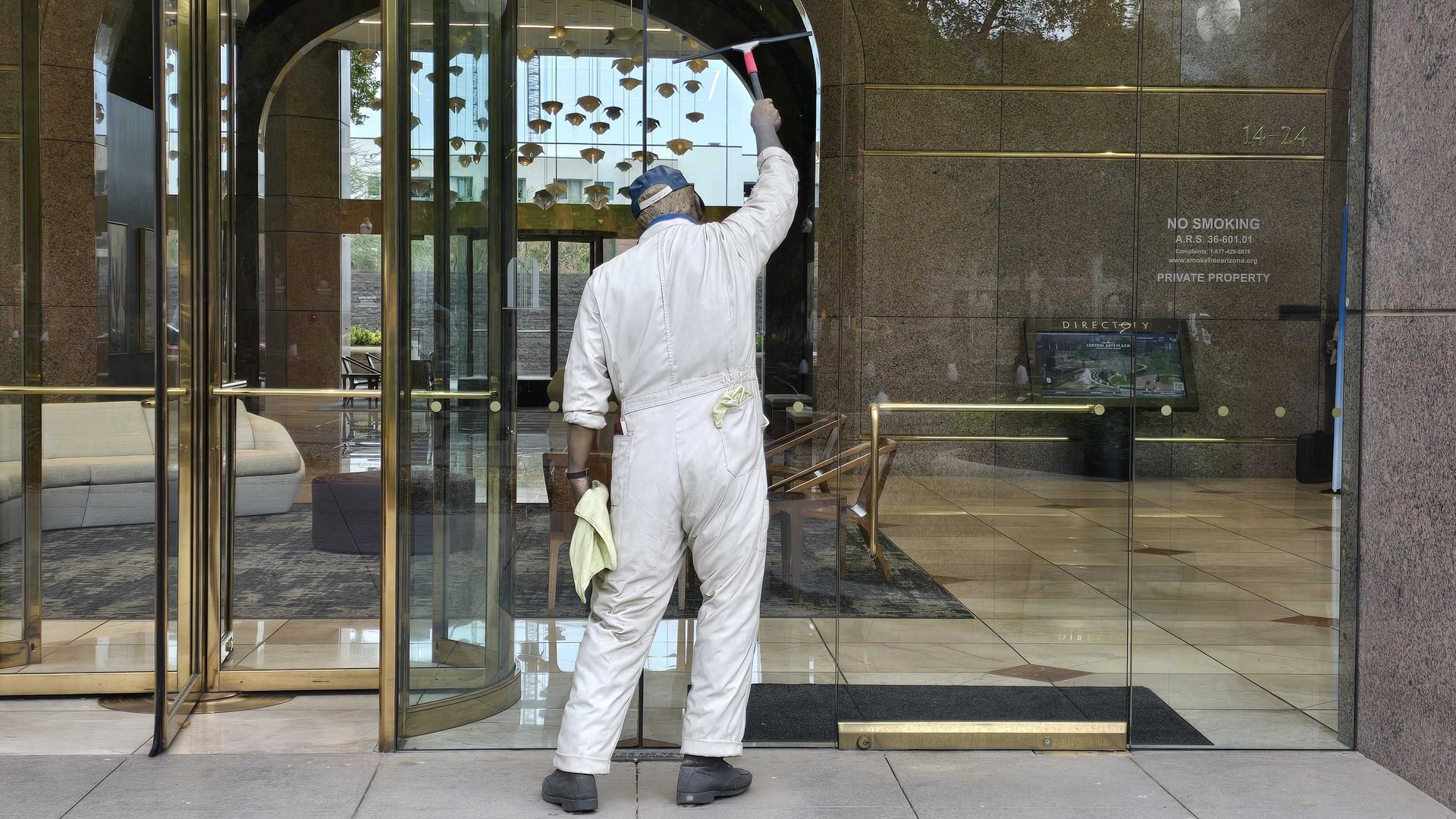 A statue of a maintenance worker in white coveralls and a blue cap wipes a glass entrance with a red-handled squeegee at a modern lobby. Gold trim, revolving doors, no-smoking sign on brown marble wall.