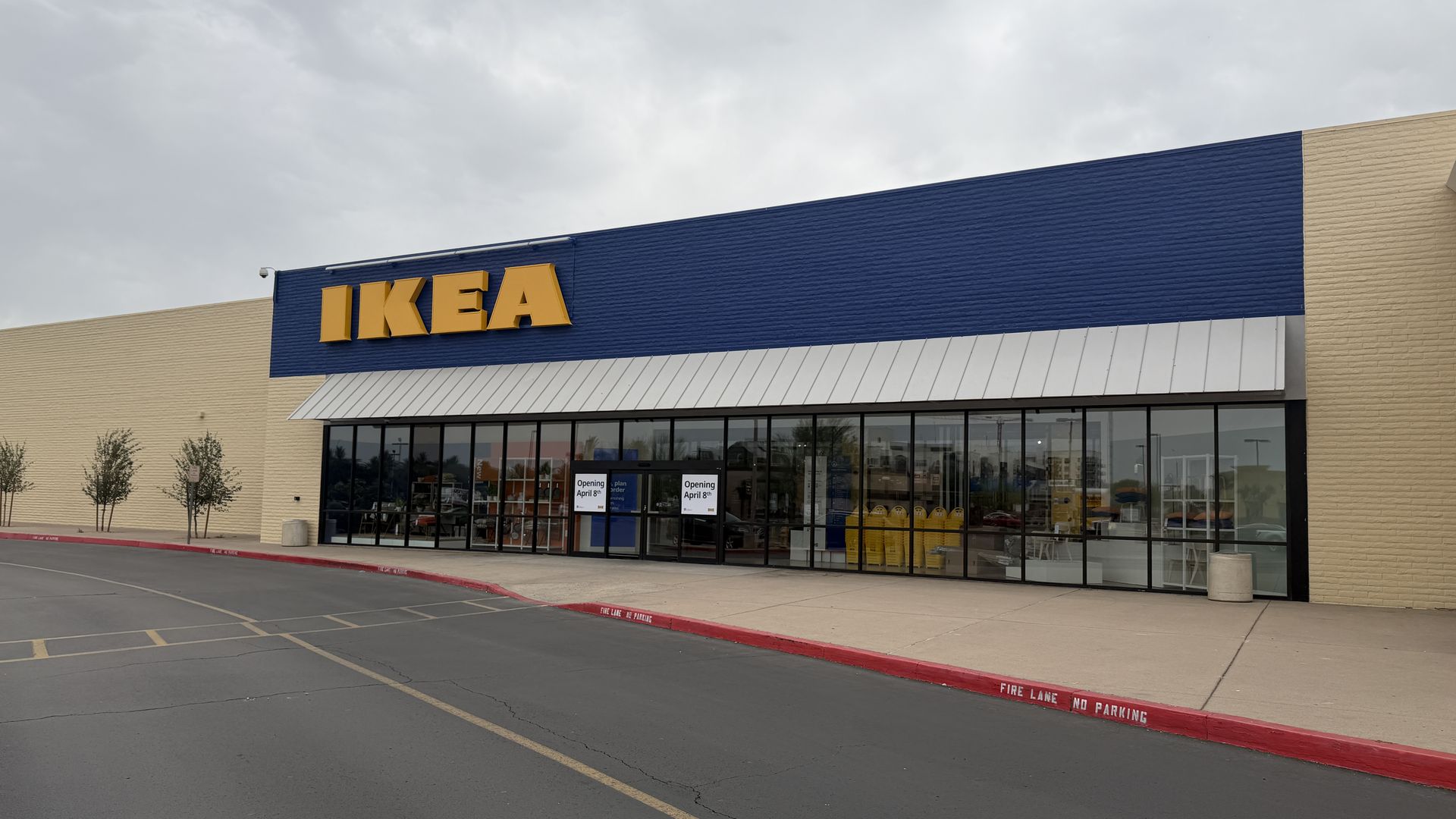 Exterior view of an IKEA store: blue wall with large yellow IKEA sign, glass storefront, beige side walls, white overhang, red curb marked FIRE LANE NO PARKING, and a cloudy sky.
