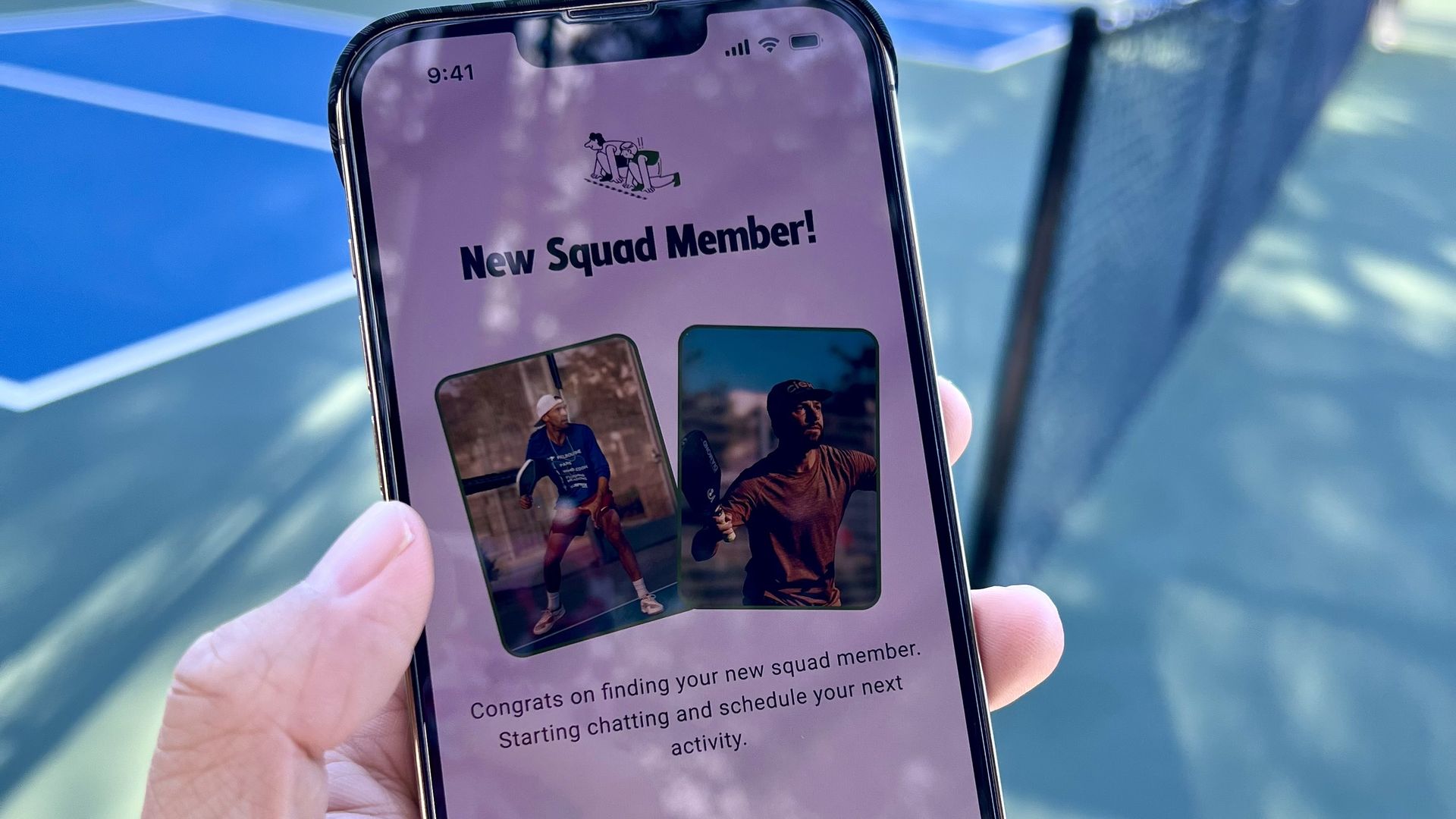 Hand holding a smartphone displaying a "New Squad Member!" screen with photos of two men and options to message or go back. The background shows blue tennis courts with a fence.