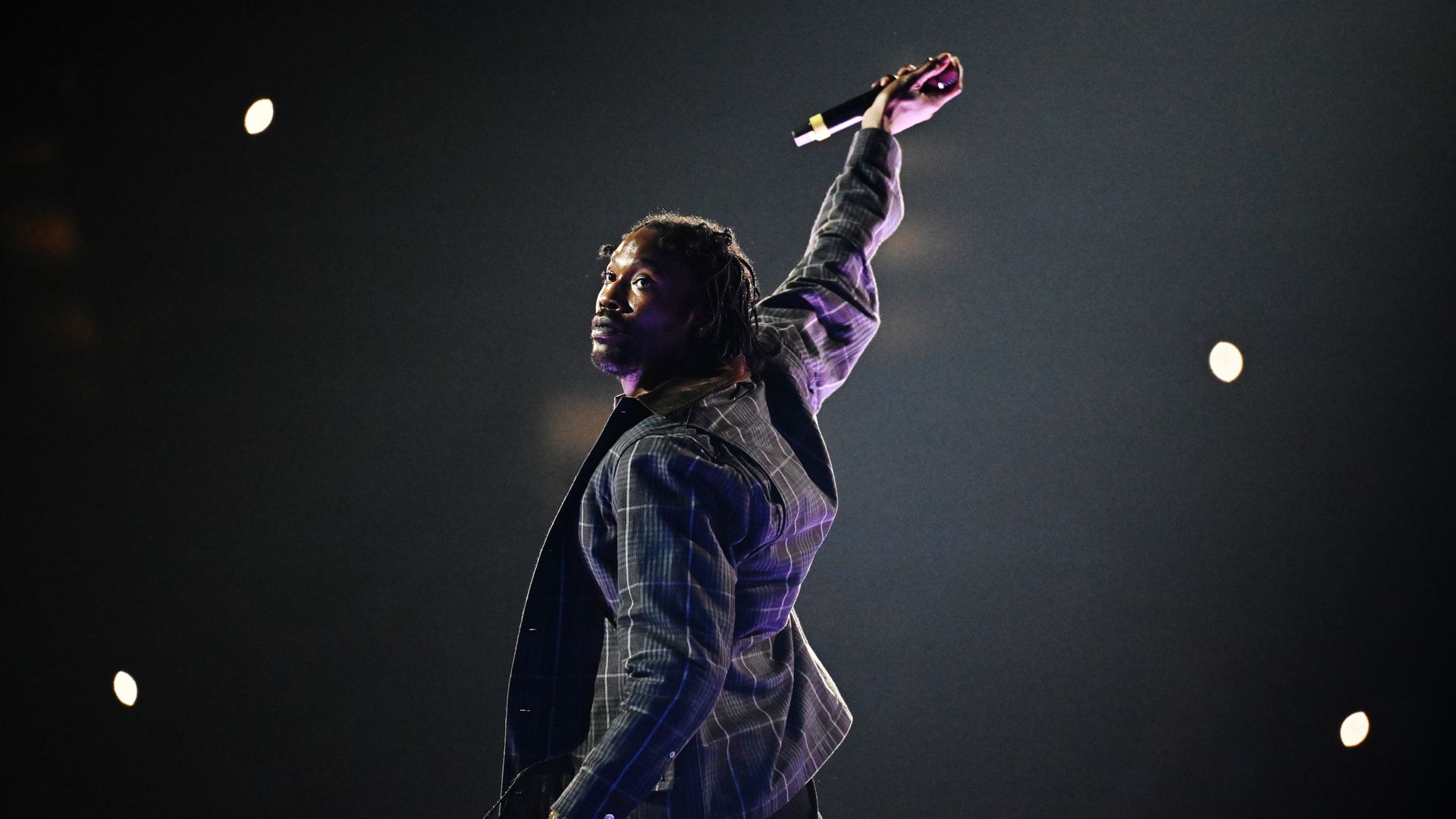 A male singer on stage raises a microphone above his head, wearing a gray plaid jacket. Purple lighting highlights his sleeve against a dark background with scattered stage lamps.