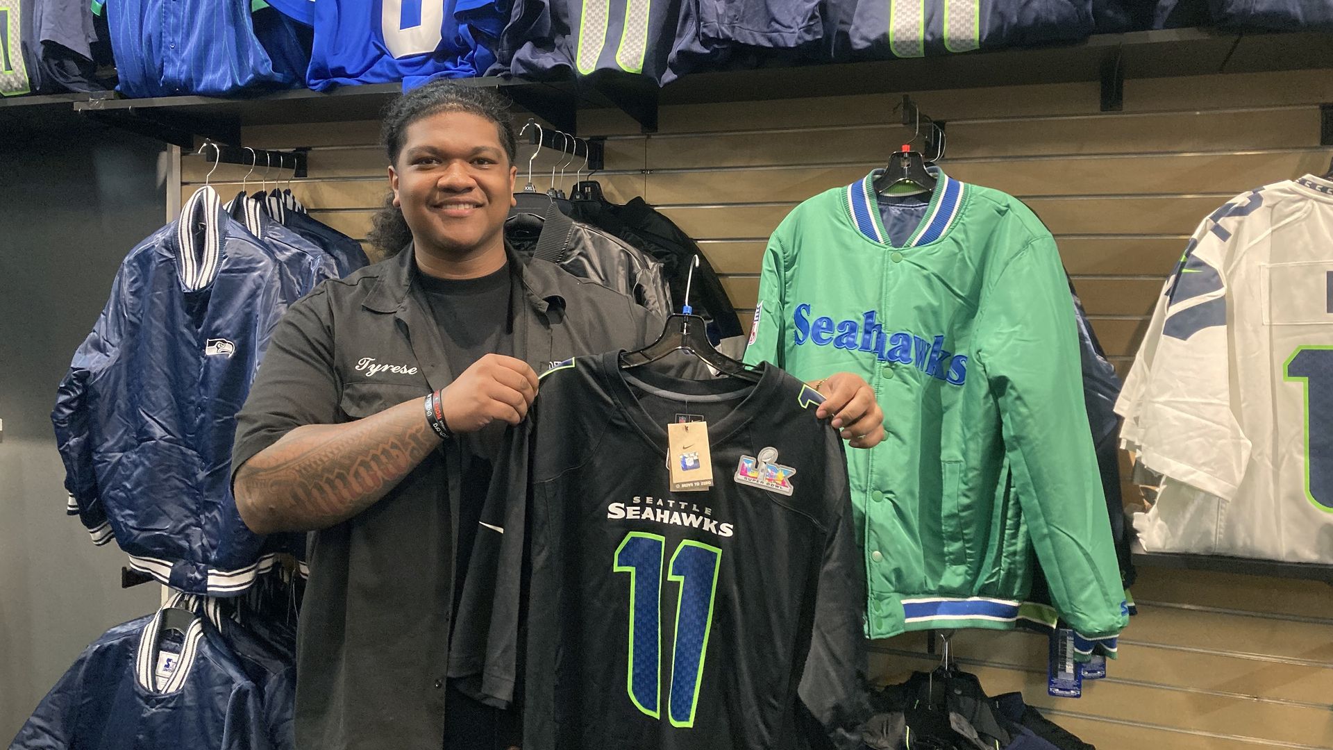 A store manager holds a Jaxon Smith-Njigba Seahawks jersey inside a shop near Lumen Field, with team apparel lining the walls behind him.