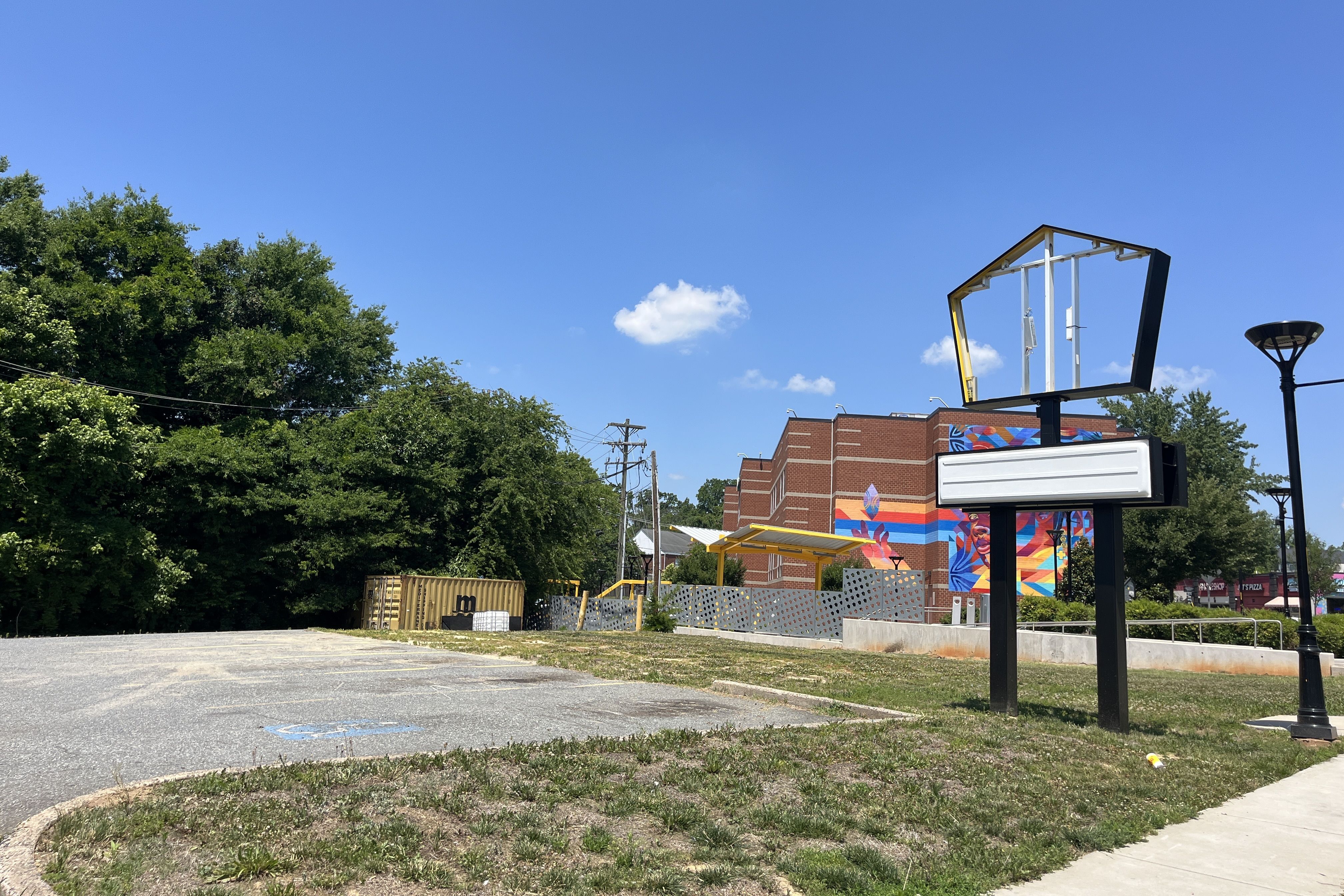 Clear blue sky above a brick building adorned with a colorful mural; in the foreground a large empty basketball hoop frame holds a blank white sign, with a street lamp nearby and green trees to the left.