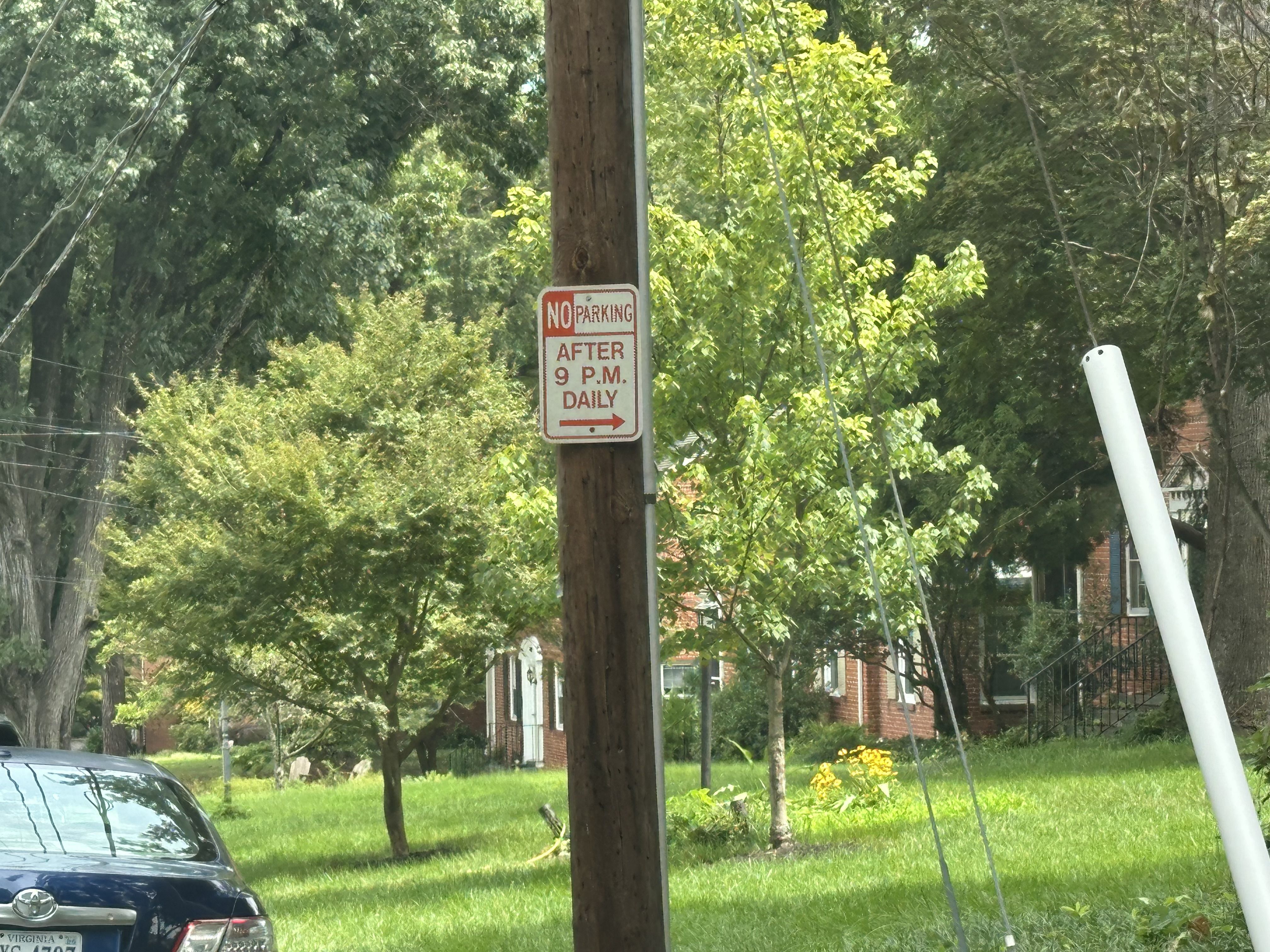 Wood utility pole with a sign reading "NO PARKING AFTER 9 P.M. DAILY" amid green trees in a residential street; a blue car sits at lower left and a white angled pole lies on the right.
