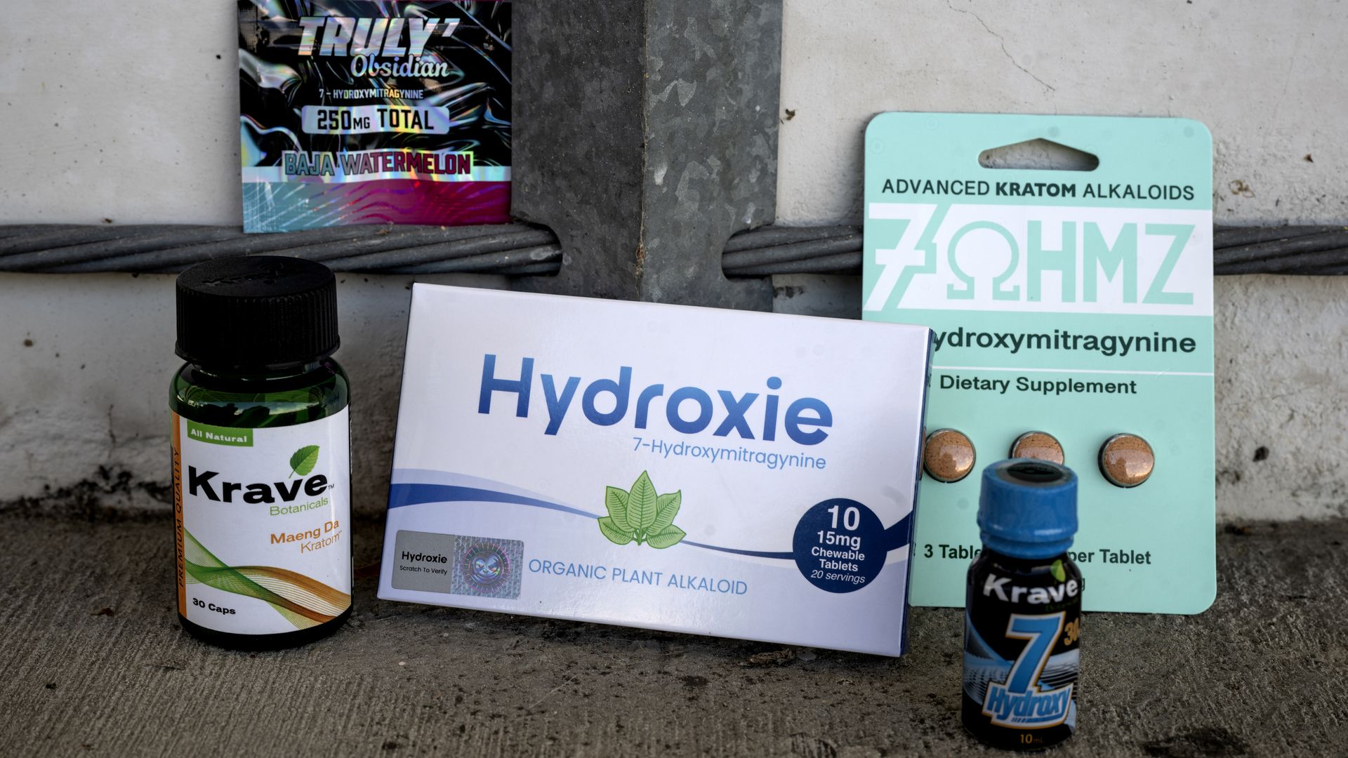 Cluttered outdoor scene of drug packaging: a large white box reading Hydroxyzine, a green Krave Botanicals bottle, a colorful foil packet labeled Baja Watermelon, and a teal Advanced Kratom Alkaloids card with pills.