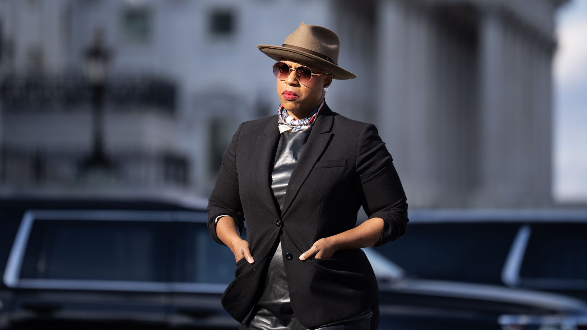 Rep. Ayanna Pressley, wearing a black suit jacket over a silver dress with a brown hat and sunglasses, stands in front of several black cars with the Capitol in the backdrop.