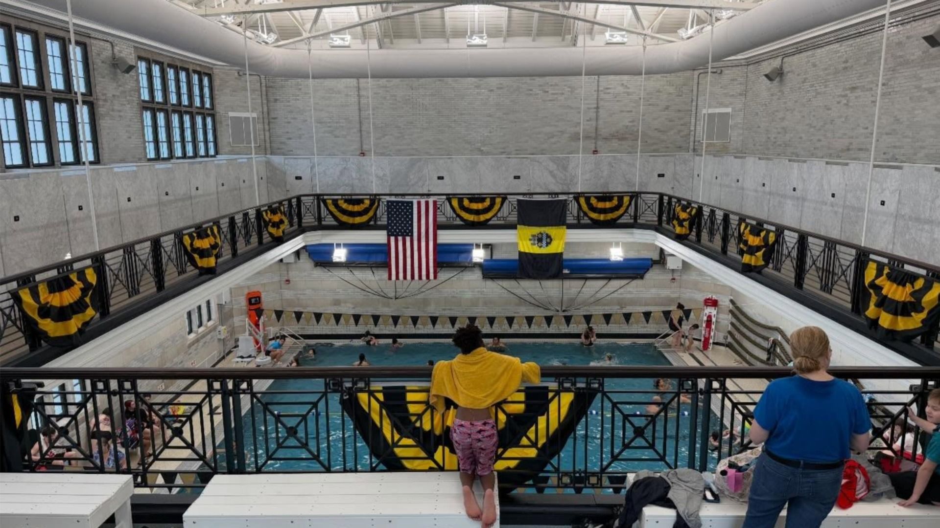 Indoor swimming pool in a brick-walled gym, lined with black-and-yellow banners. An American flag and a black-and-yellow flag hang over the pool. A person with a yellow towel stands at the railing watching swimmers.