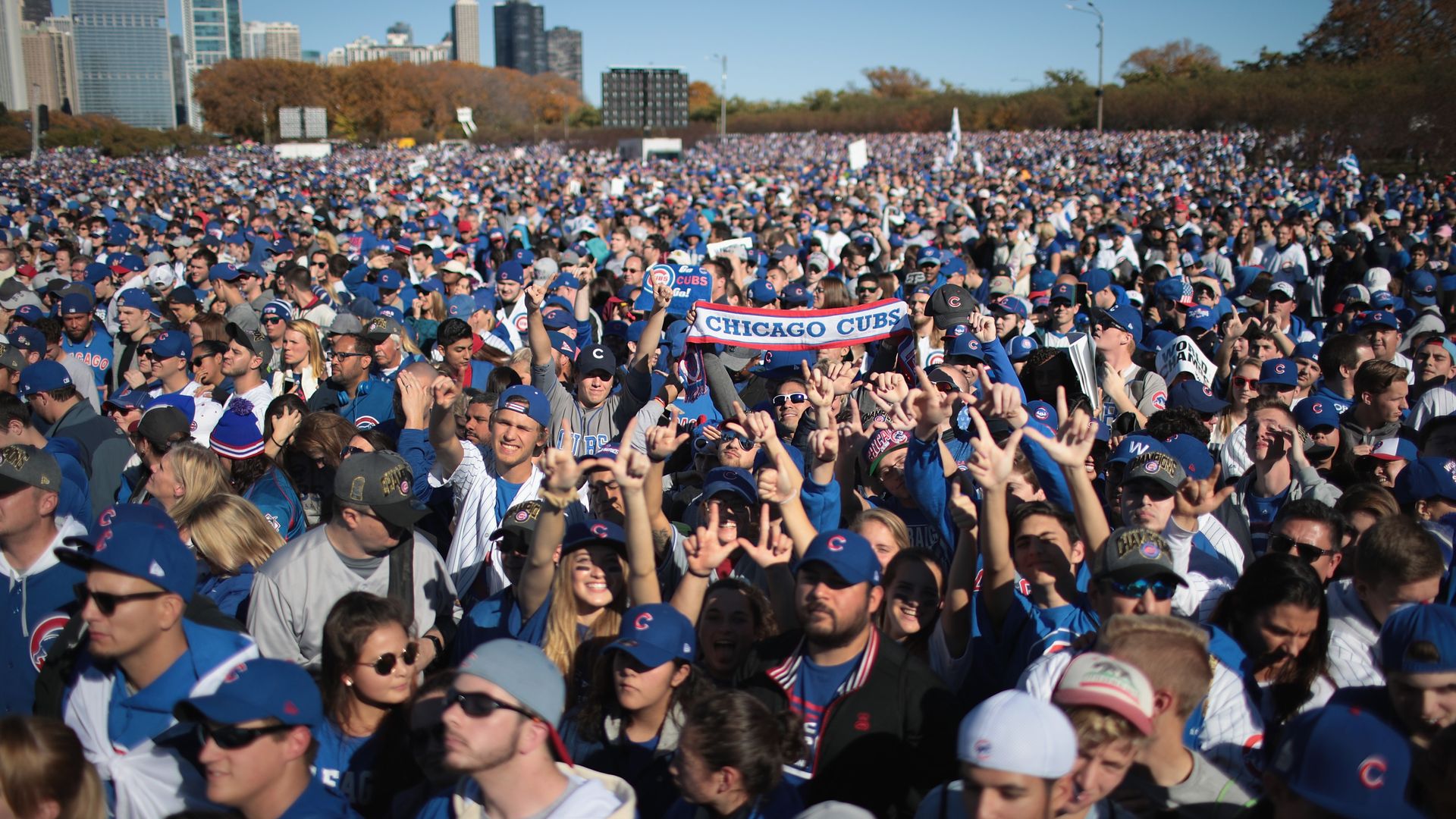 Large crowd wearing blue Chicago Cubs gear, hats, and jerseys gathers outdoors in a sunny park with city skyscrapers in the background, showing support with signs and scarves.