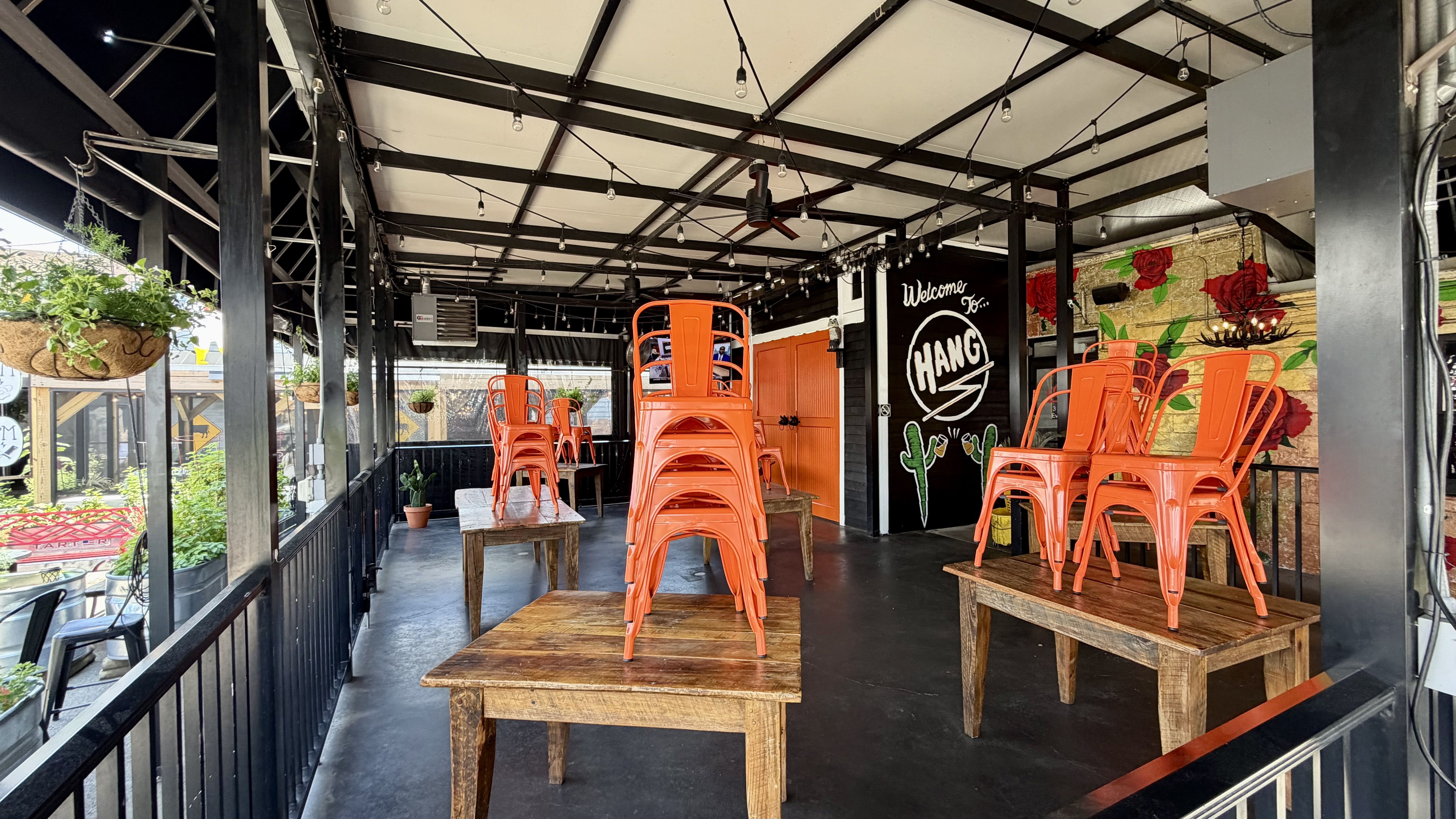 Open-air industrial dining space with bright orange stacked metal chairs on wooden platforms, black steel beams and string lights, hanging plants, and a mural wall with red roses.