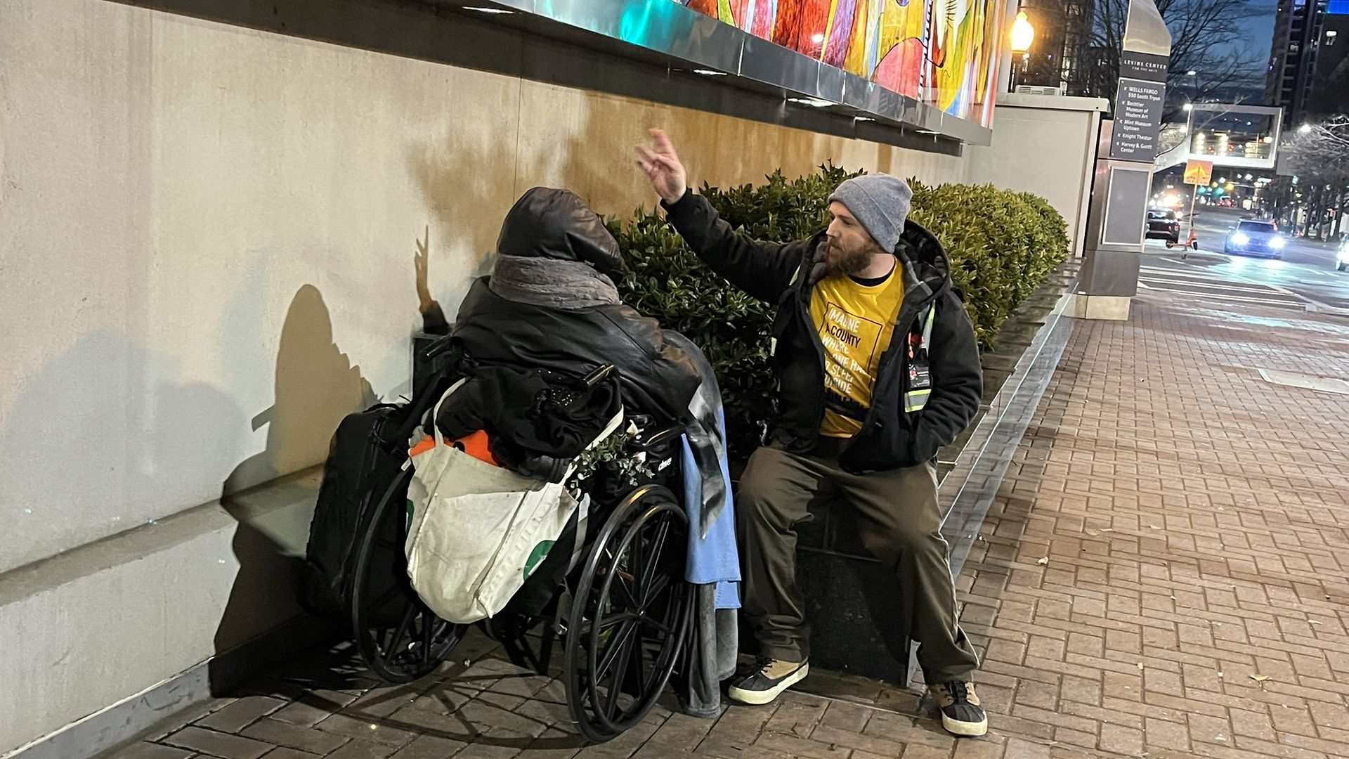 Night city sidewalk: a bearded man in a gray beanie and yellow shirt raises a hand toward a person in a wheelchair piled with bags; a bright mural towers above, streetlights and cars in background.
