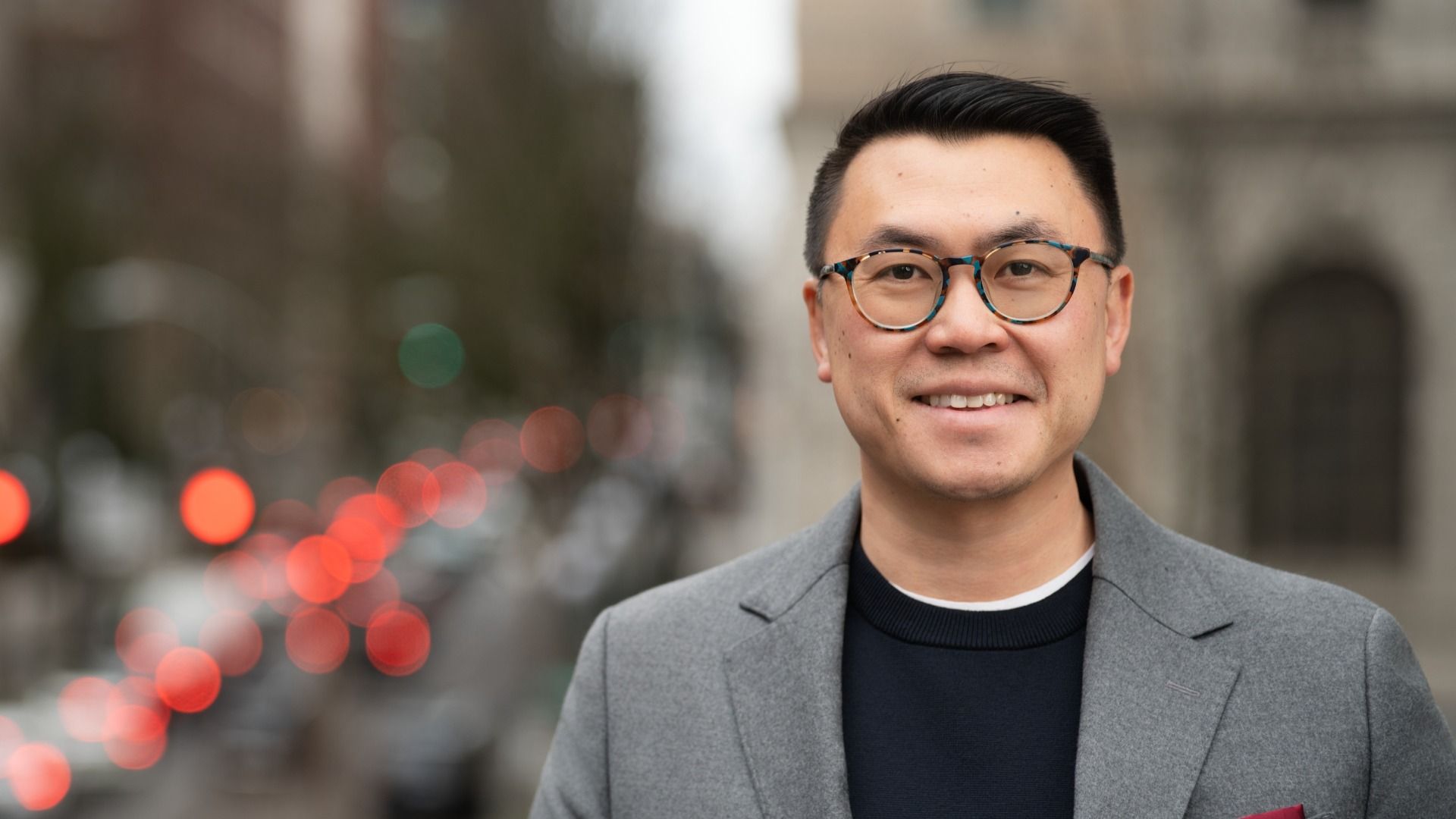Portrait of a man with round tortoiseshell glasses, gray coat over a dark sweater, smiling at the camera on a city street with blurred red brake lights.