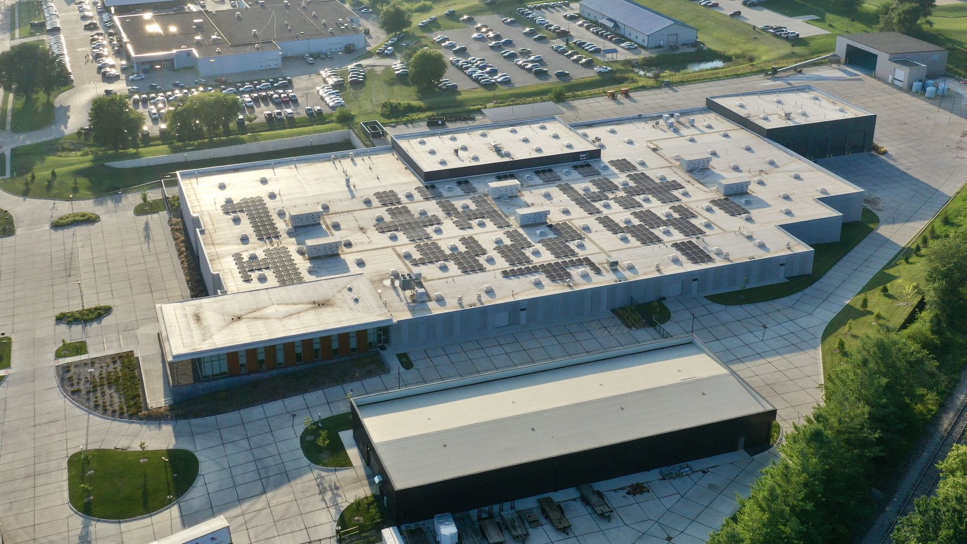 Aerial view of a large industrial building with solar panels on its flat roof, surrounded by parking lots, smaller buildings, green trees, and paved roads under sunlight.