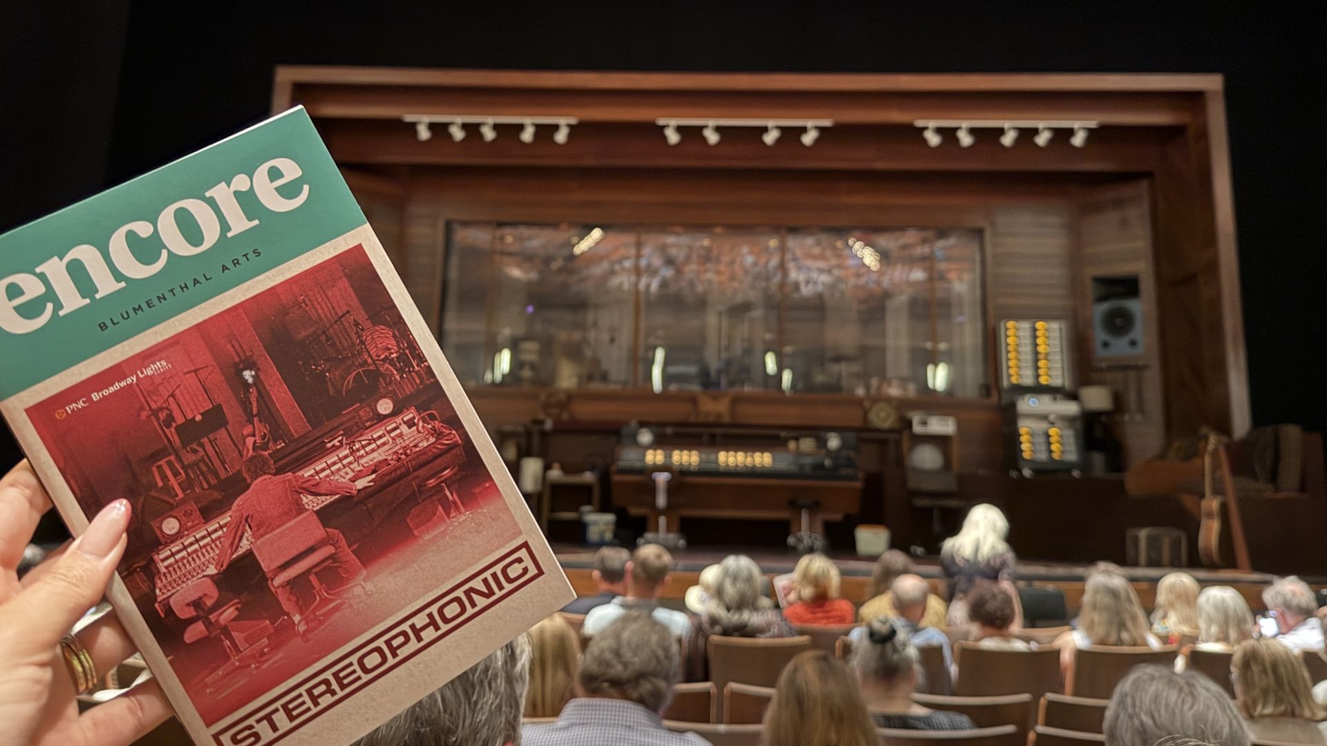 Foreground shows a hand holding a teal magazine cover reading " Encore" with a red-tinted photo and "STEREOPHONIC" at the bottom; a wooden theater stage and seated audience in the background.