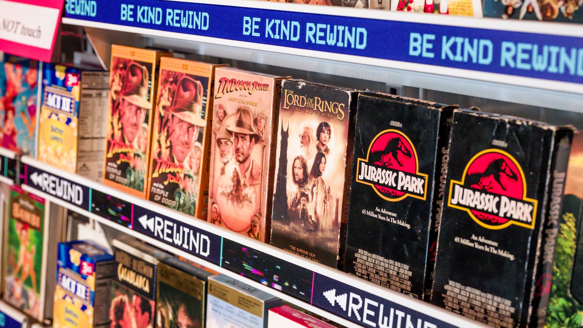 A photo of VHS tapes like Lord of the Rings and Jurassic Park lined up on a shelf below a sign that says Be Kind, Rewind. 
