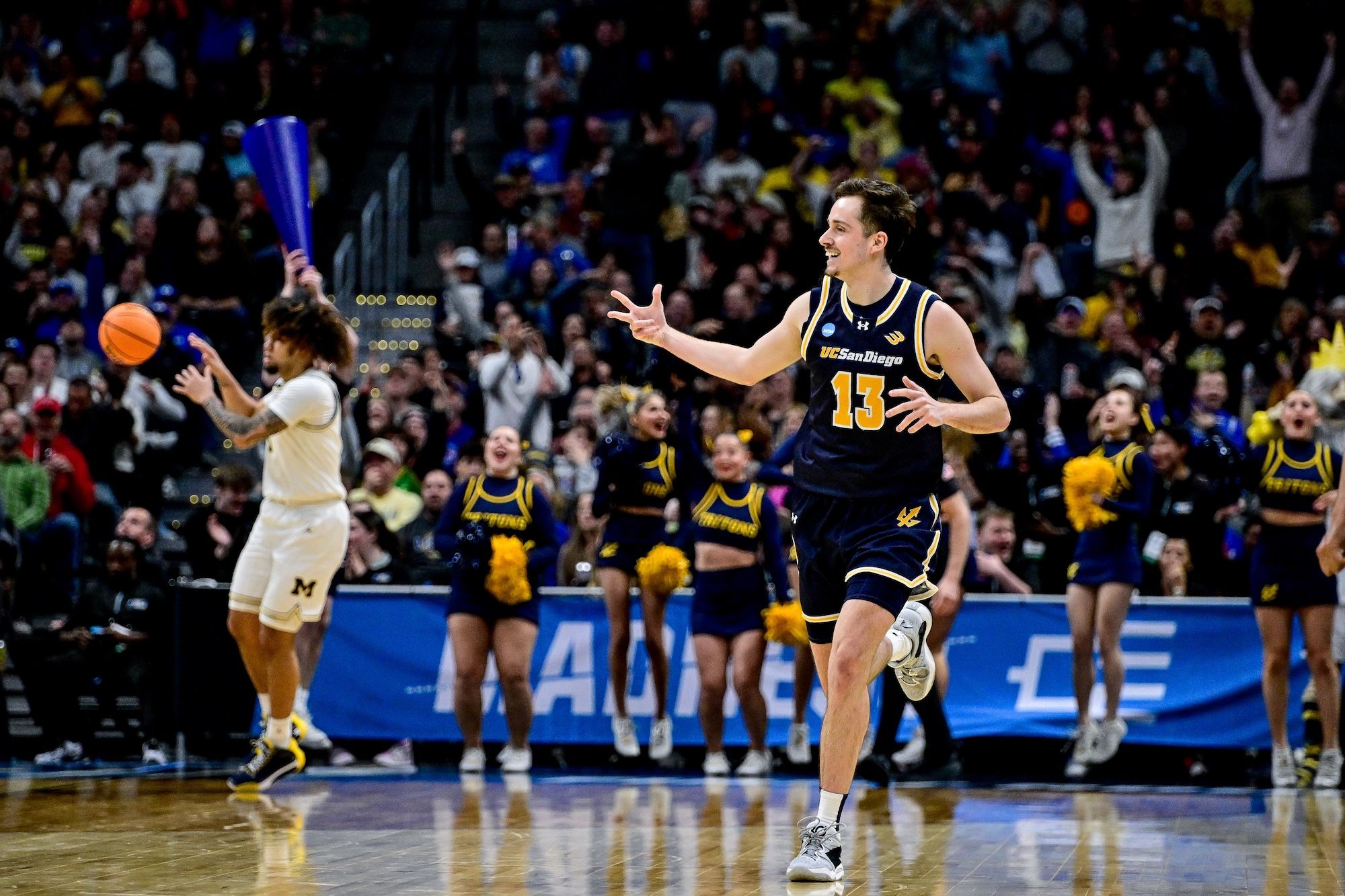 A UCSD men's basketball player holds up 3 fingers to the crowd while running down the court after making a shot.
