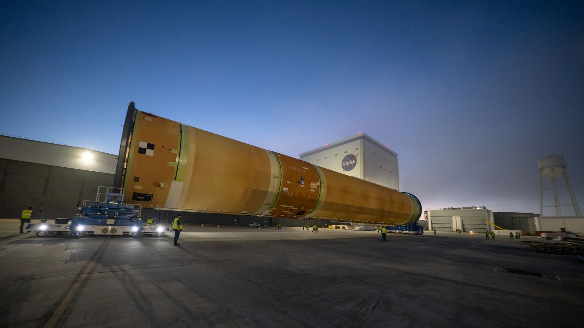 Nighttime at a NASA facility: a large orange rocket stage lies on its side on the tarmac, supported by equipment. Ground crew in hi-vis vests observe, with a NASA building in the background.