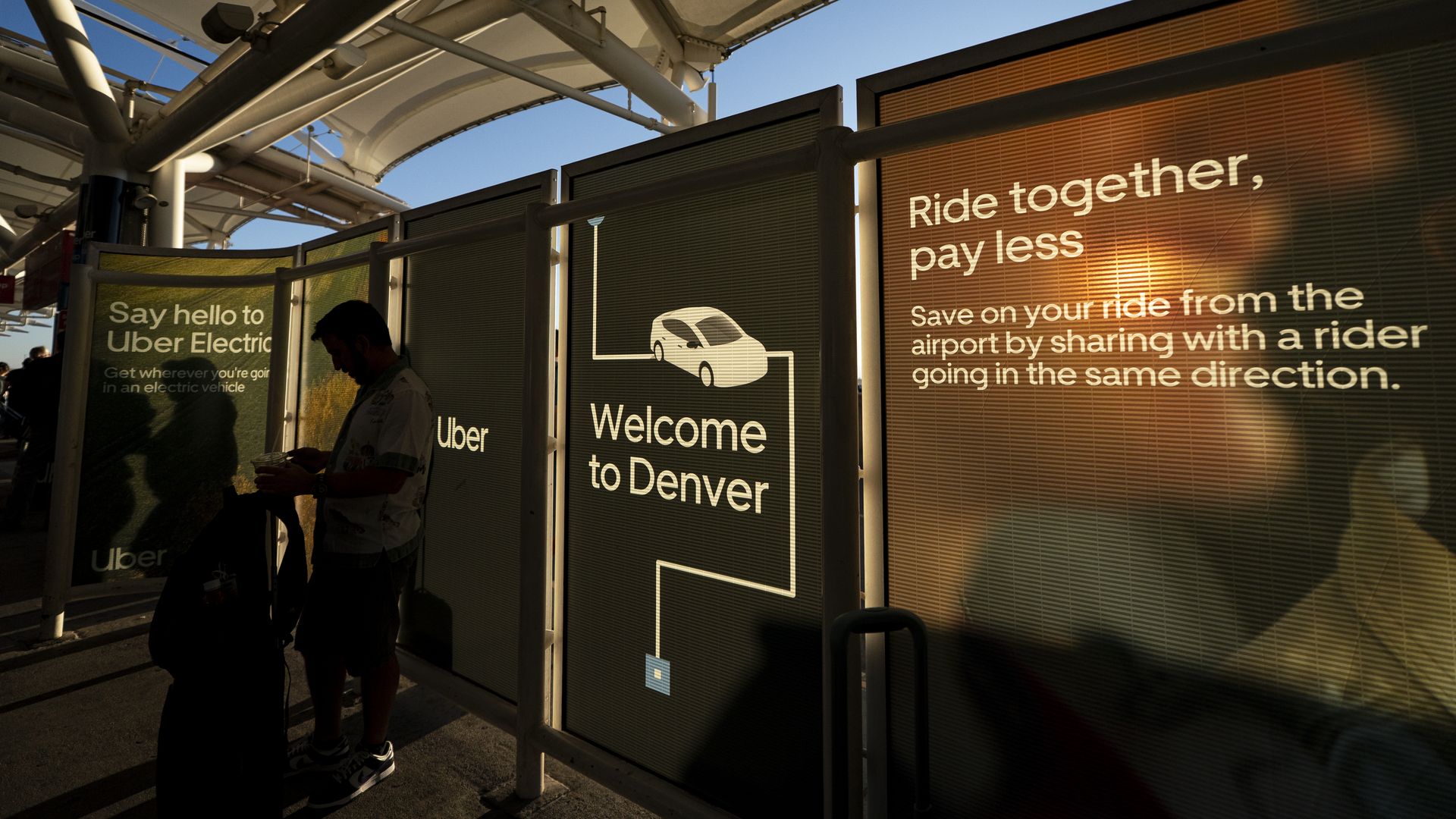 Sunlit transit walkway with white metal framework and large glass panels displaying "Ride together, pay less" and "Welcome to Denver"; a person silhouette stands left, blue sky above.