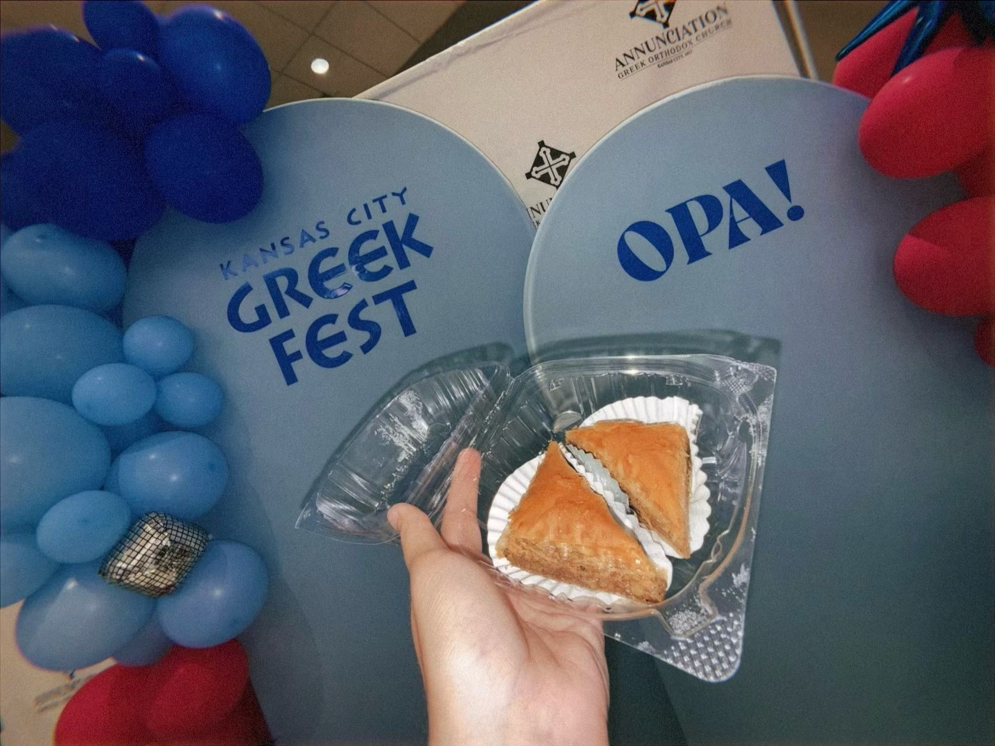Hand holding plastic container with two pieces of baklava in front of blue balloons with text "Kansas City Greek Fest" and "OPA!", and a sign for Annunciation Greek Orthodox Church.