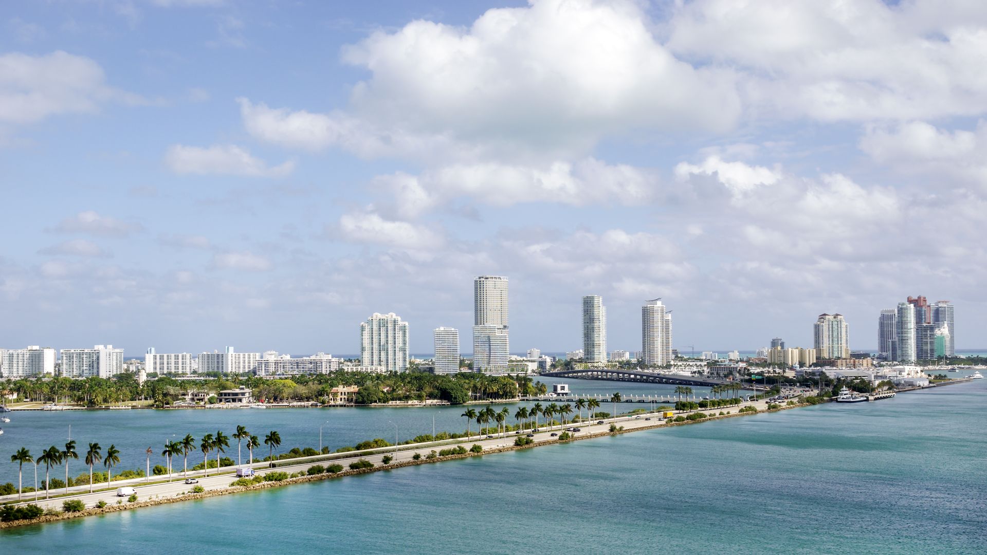 aerial view of a palm tree-lined road surrounding by clear blue water on either side and the miami skyline in the back