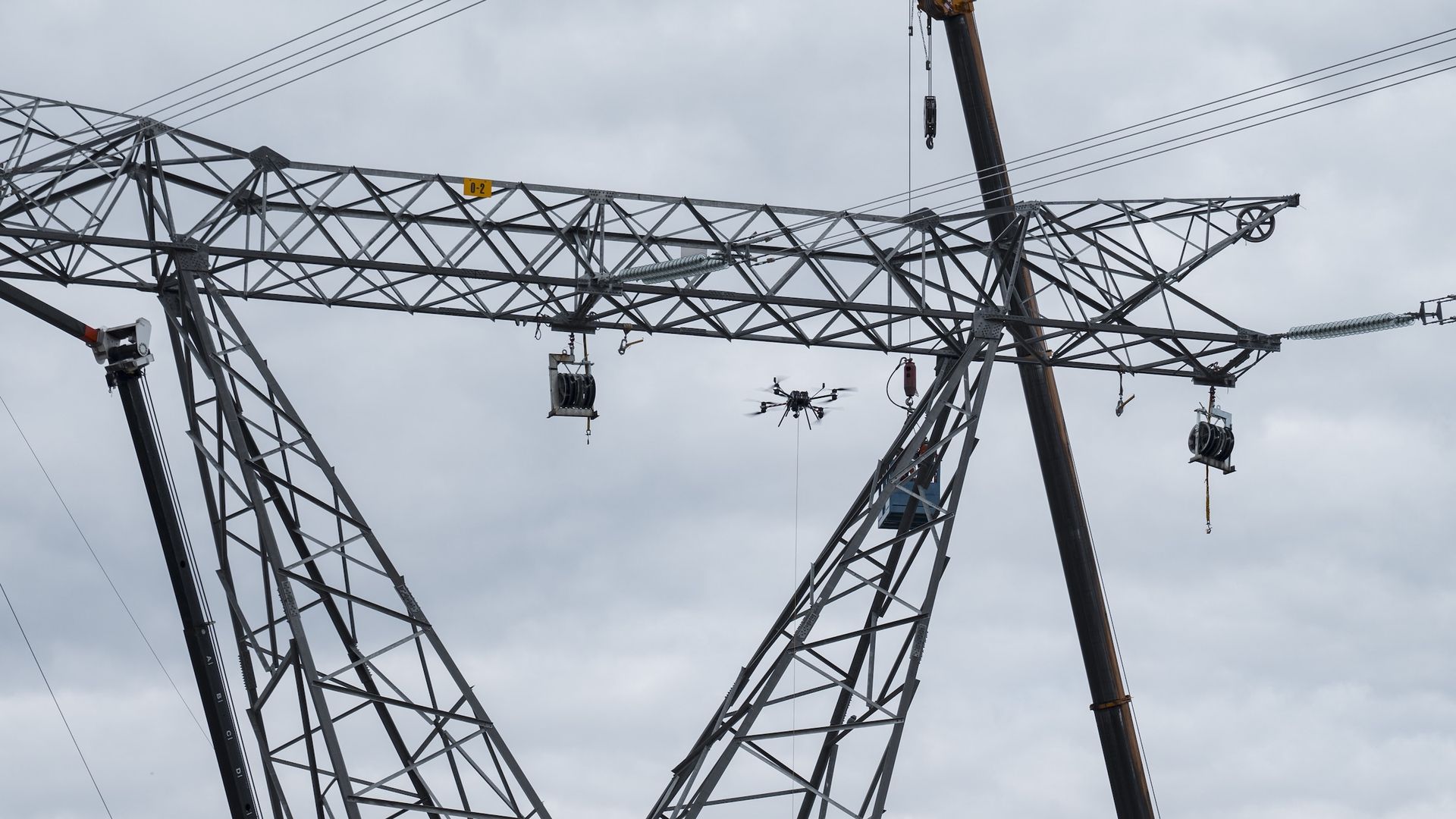 Drone inspecting or working on a large metal electrical transmission tower with overcast sky background and cranes nearby.