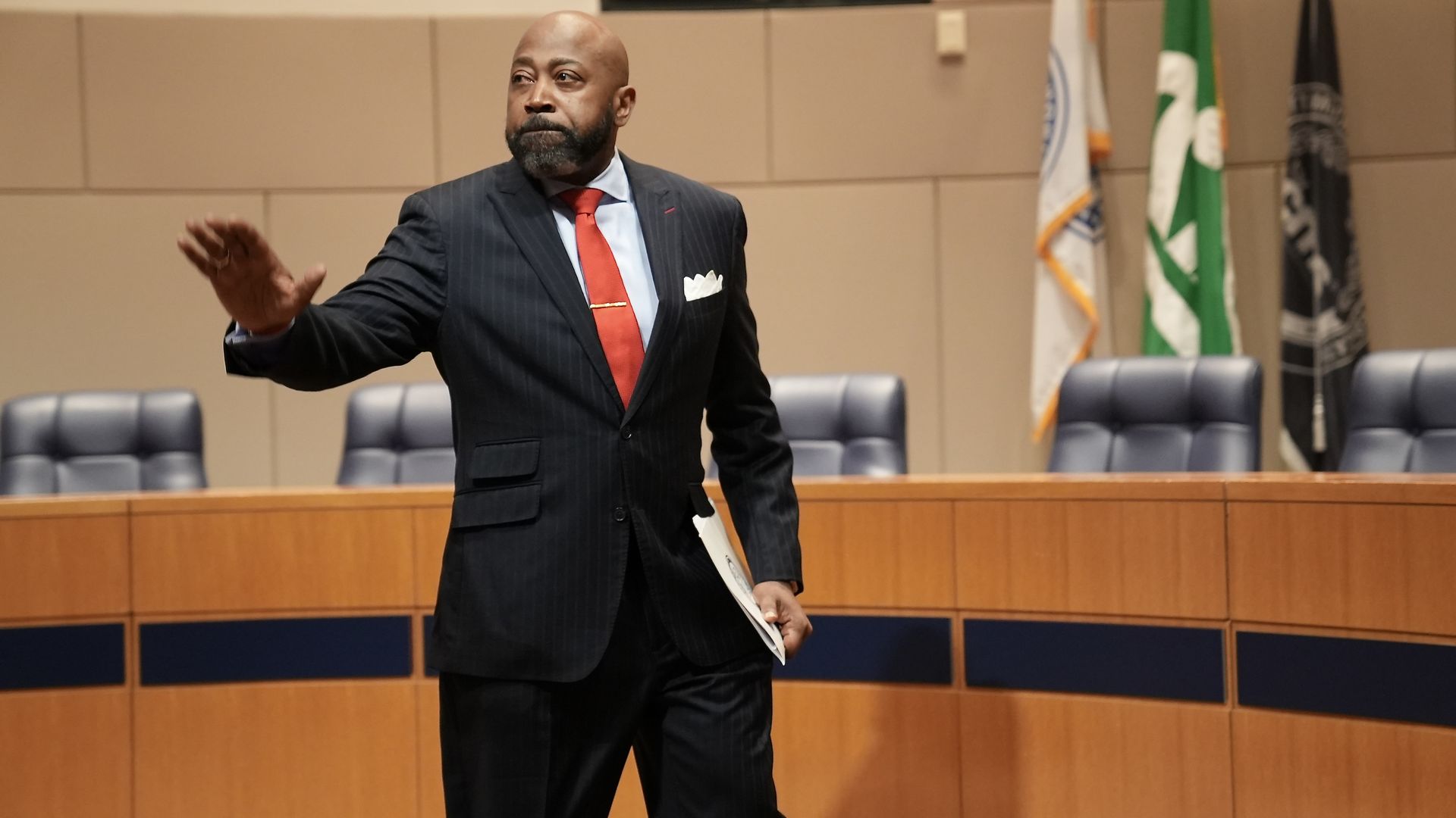 Man in a dark pinstripe suit with a red tie, holding papers and gesturing with his hand, standing in a council chamber with blue chairs and flags in the background.